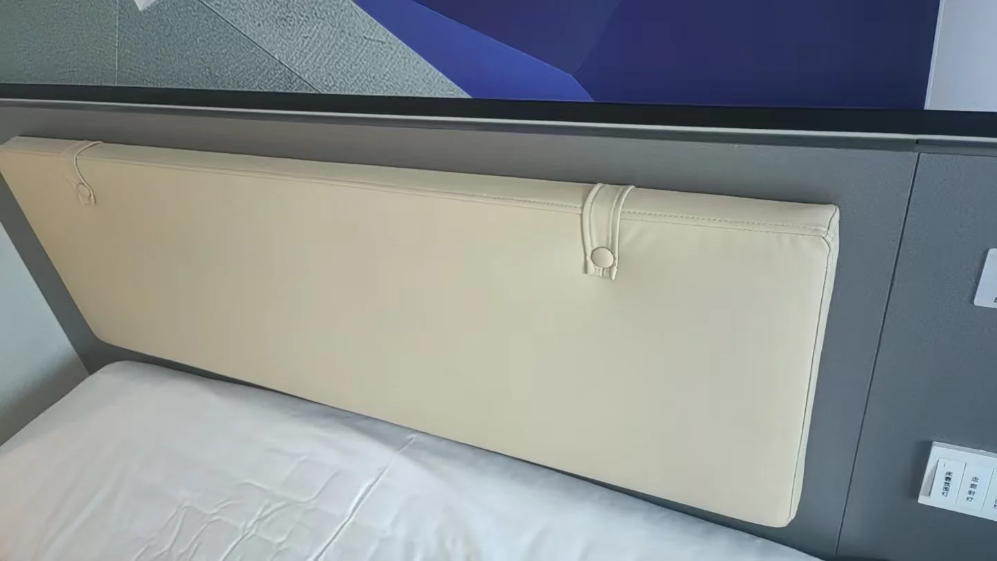 Hospital or hotel bed with a padded beige headboard attached to a grey wall.