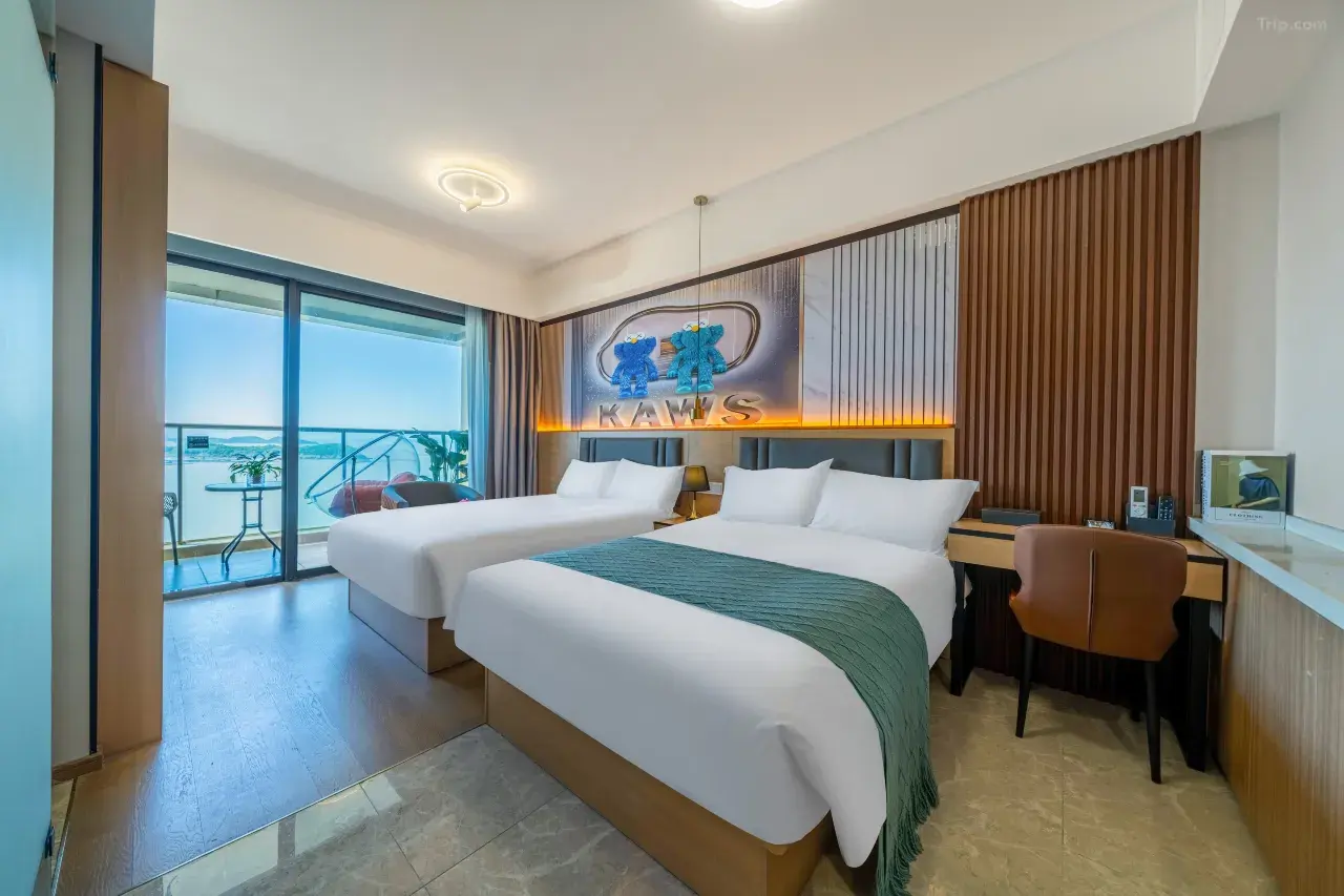 Hotel room with two beds, a balcony with outdoor seating, a wall decor featuring two teddy bears and the word 'KAWS' illuminated, and a desk with a chair and electronic devices.