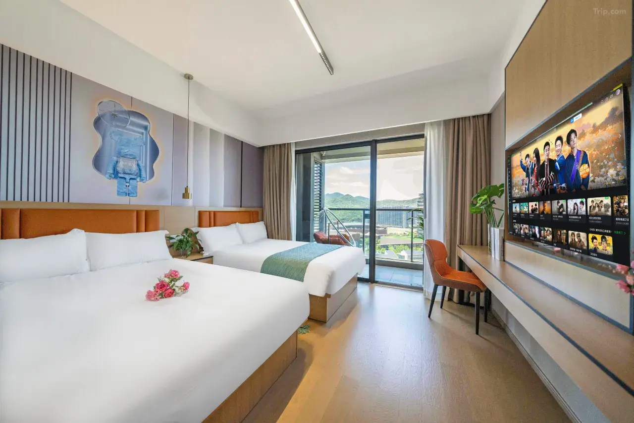 A modern hotel room with two beds, a balcony with a view of mountains and city buildings, a large flat-screen TV, and a desk with an orange chair.