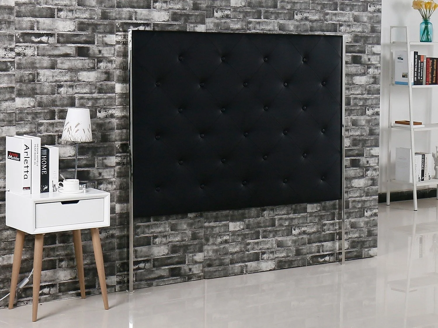 Interior of a modern bedroom with exposed brick wall, a large black tufted headboard, white side table with books, lamp, and teacup, and a white bookshelf with decorative items and books.
