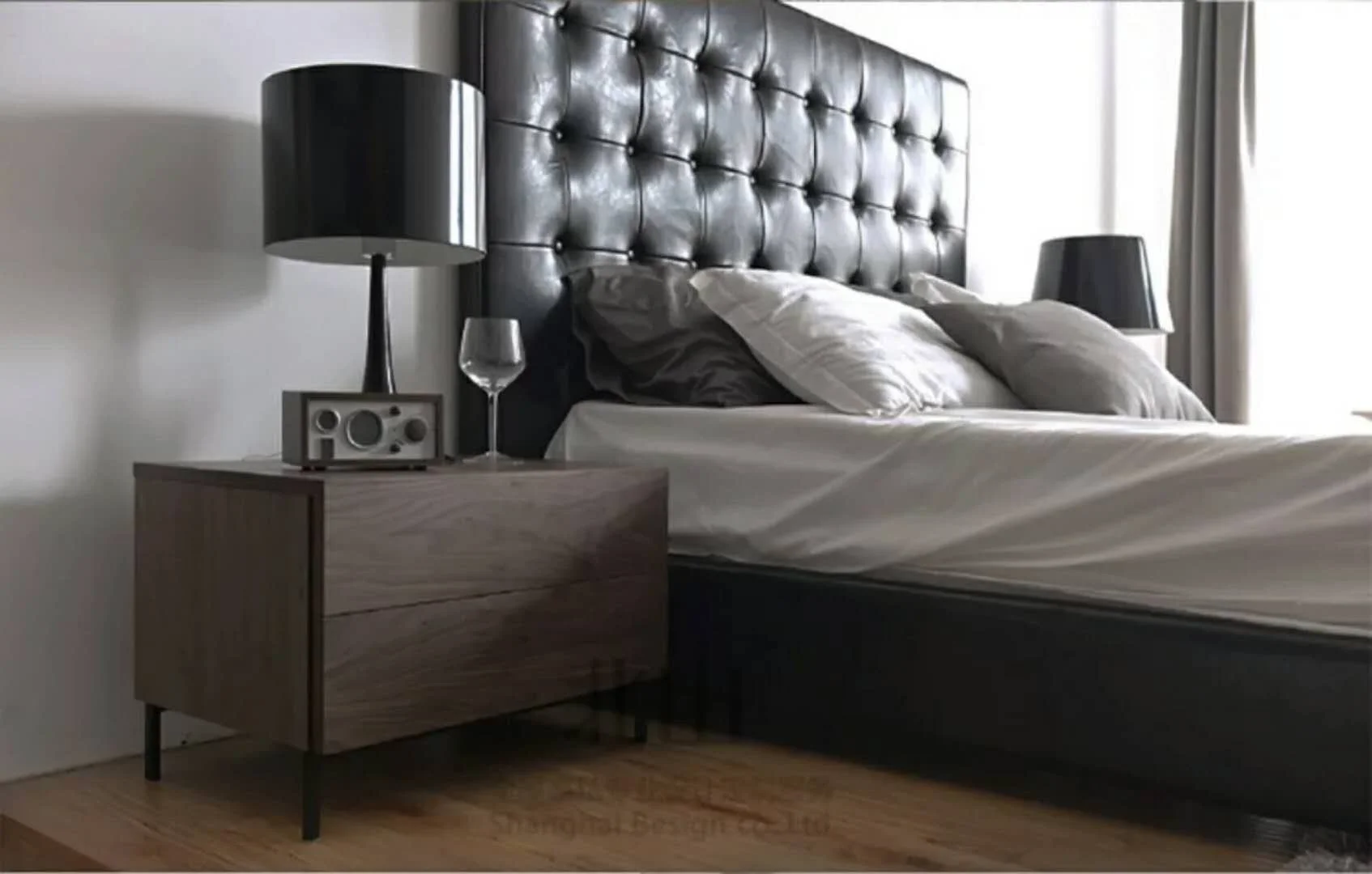 A modern bedroom with a dark upholstered headboard, white bedding, and a wooden nightstand with black lamps, a wine glass, and a clock radio.