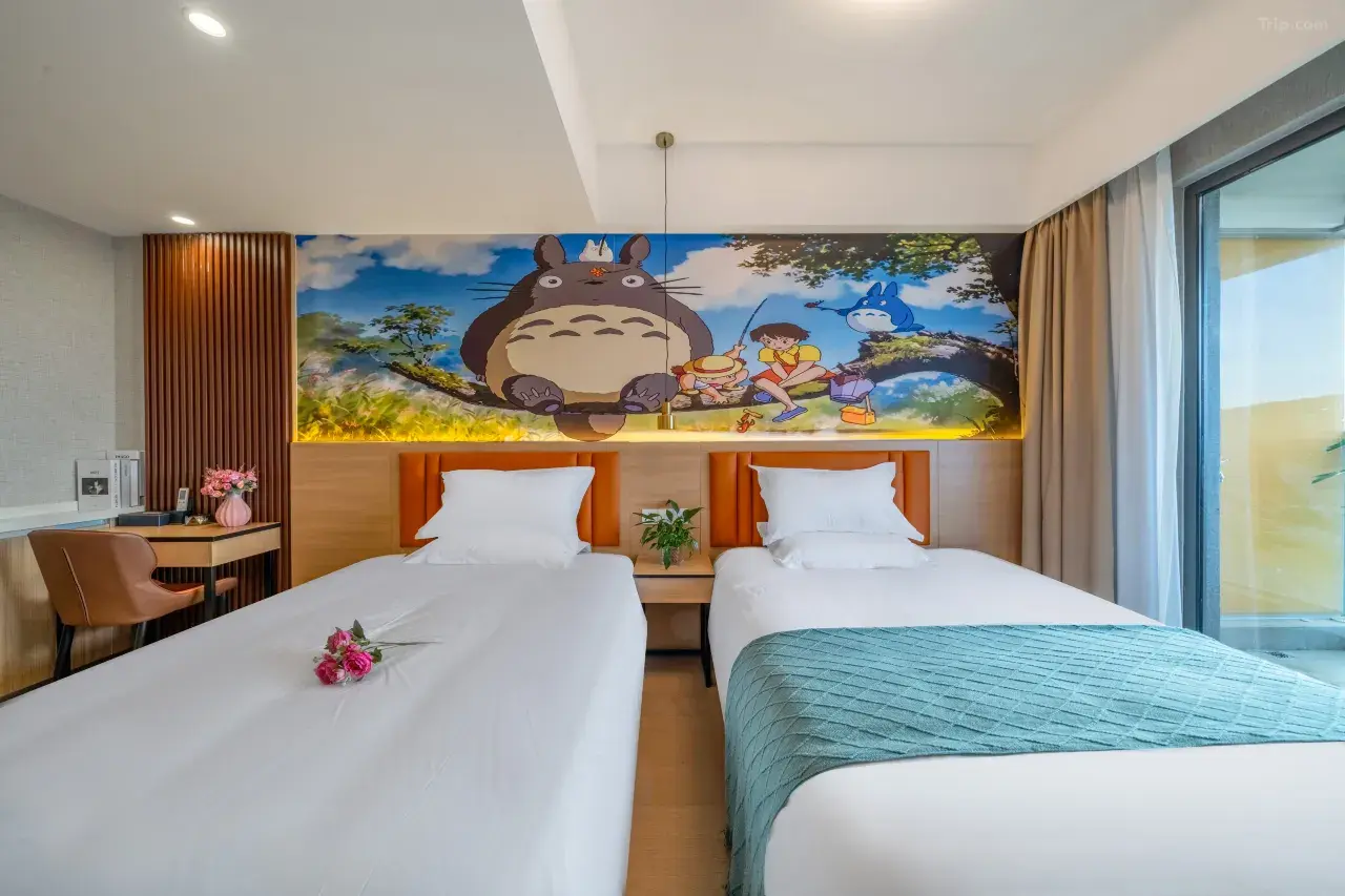 Hotel room with two beds featuring a Totoro mural on the wall above, decorated with flowers on the beds and a small side table with a plant, near a large window with curtains.