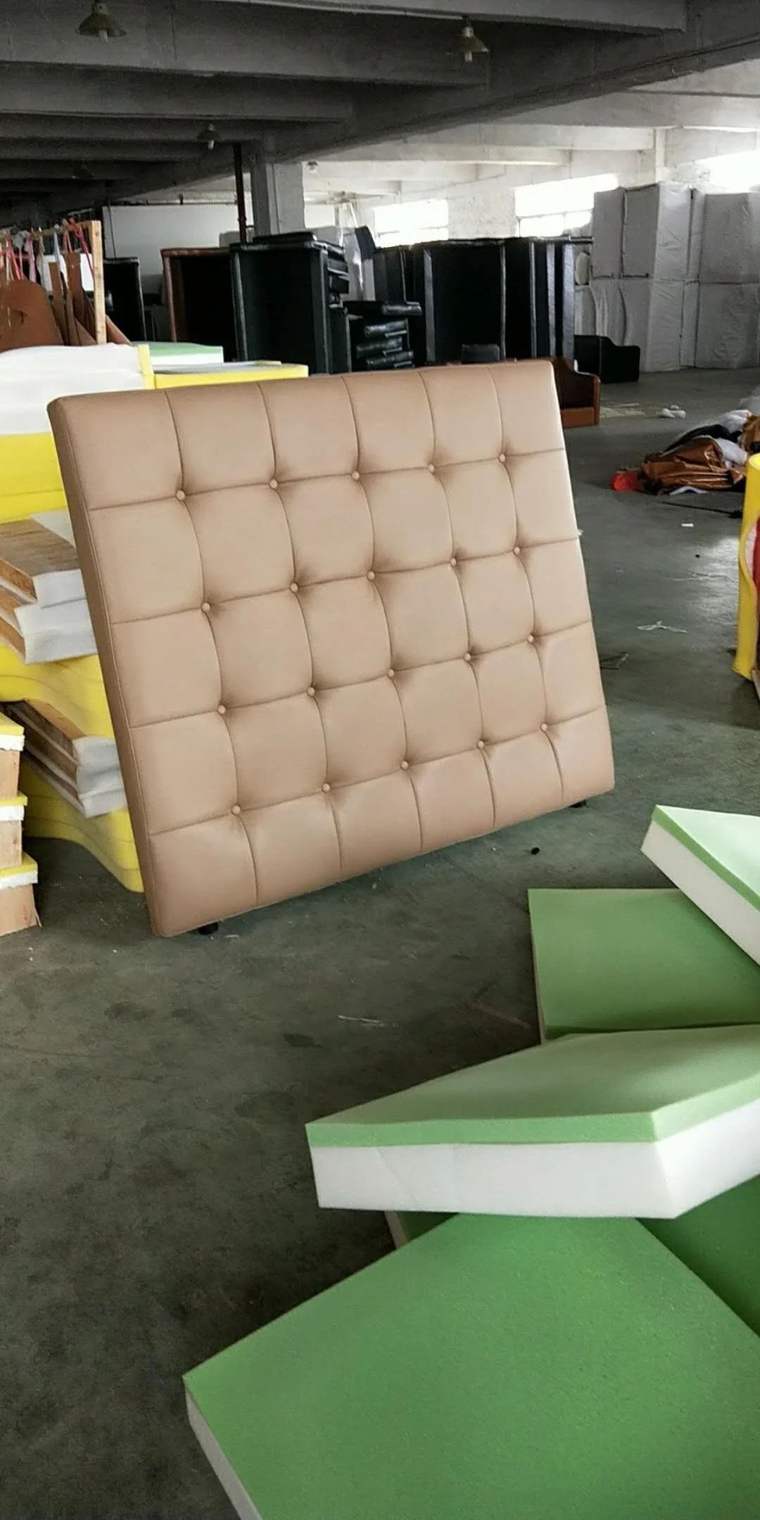 A beige, quilted, tufted headboard leaning against a wall in a warehouse or storage area.