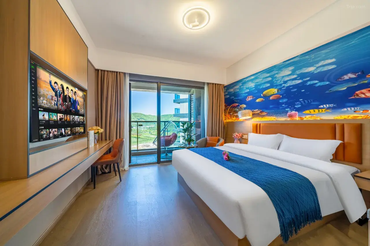 A hotel room with a large bed that has white linens and a dark blue throw blanket, a wall mural of an underwater scene with fish, a balcony with a chair and plants, a flat-screen TV on a wooden wall, and a view of green hills outside.