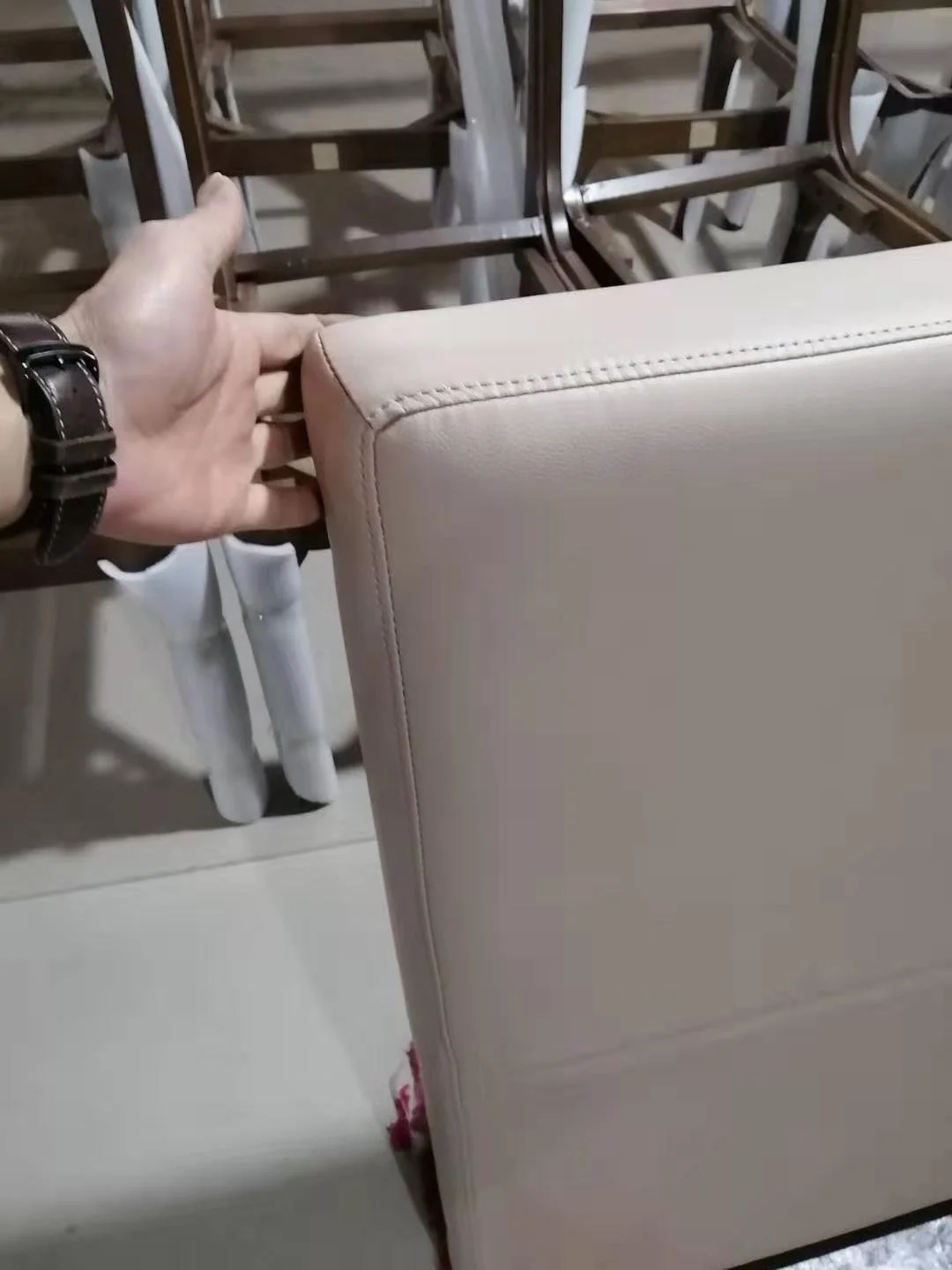 Person inspecting the back corner of a beige upholstered chair with wooden chairs stacked in the background.