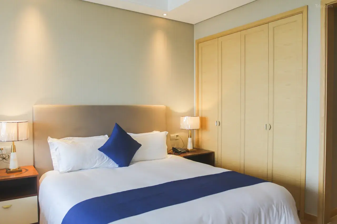 Hotel room with a bed, beige headboard, white bedding with a blue runner, a single blue pillow, two side tables with lamps, and a closet with double sliding doors.