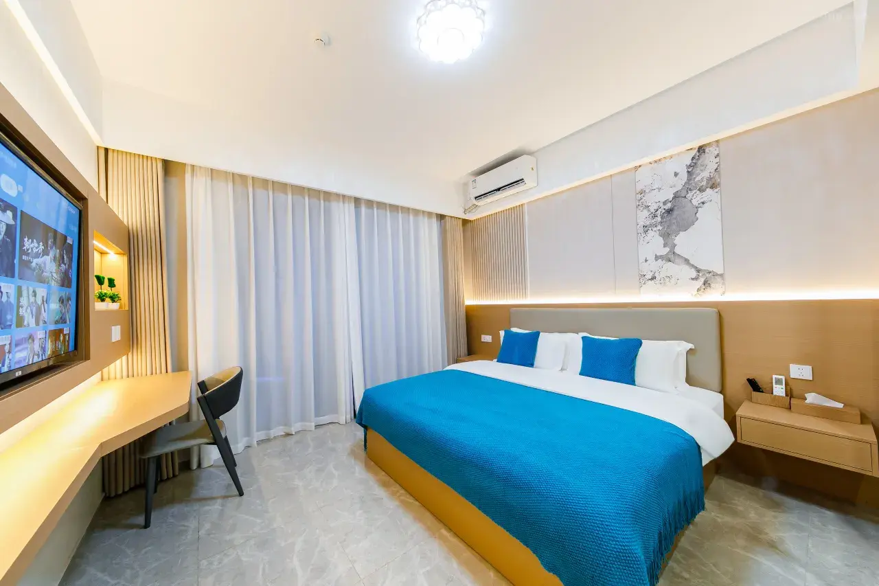 A modern hotel room with a large bed covered in a blue bedspread, four pillows, and a beige headboard. There is a wall-mounted TV on the left, a desk with a chair underneath, and curtains covering a window on the far side. The room has neutral-colore