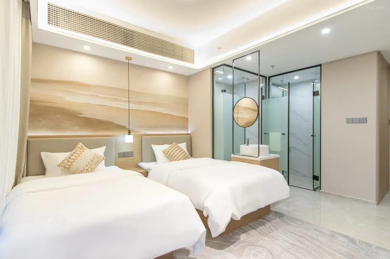 Modern hotel room with two twin beds, a shared bedside table, pillows, a pendant light, and a glass-enclosed bathroom with a sink and shower.