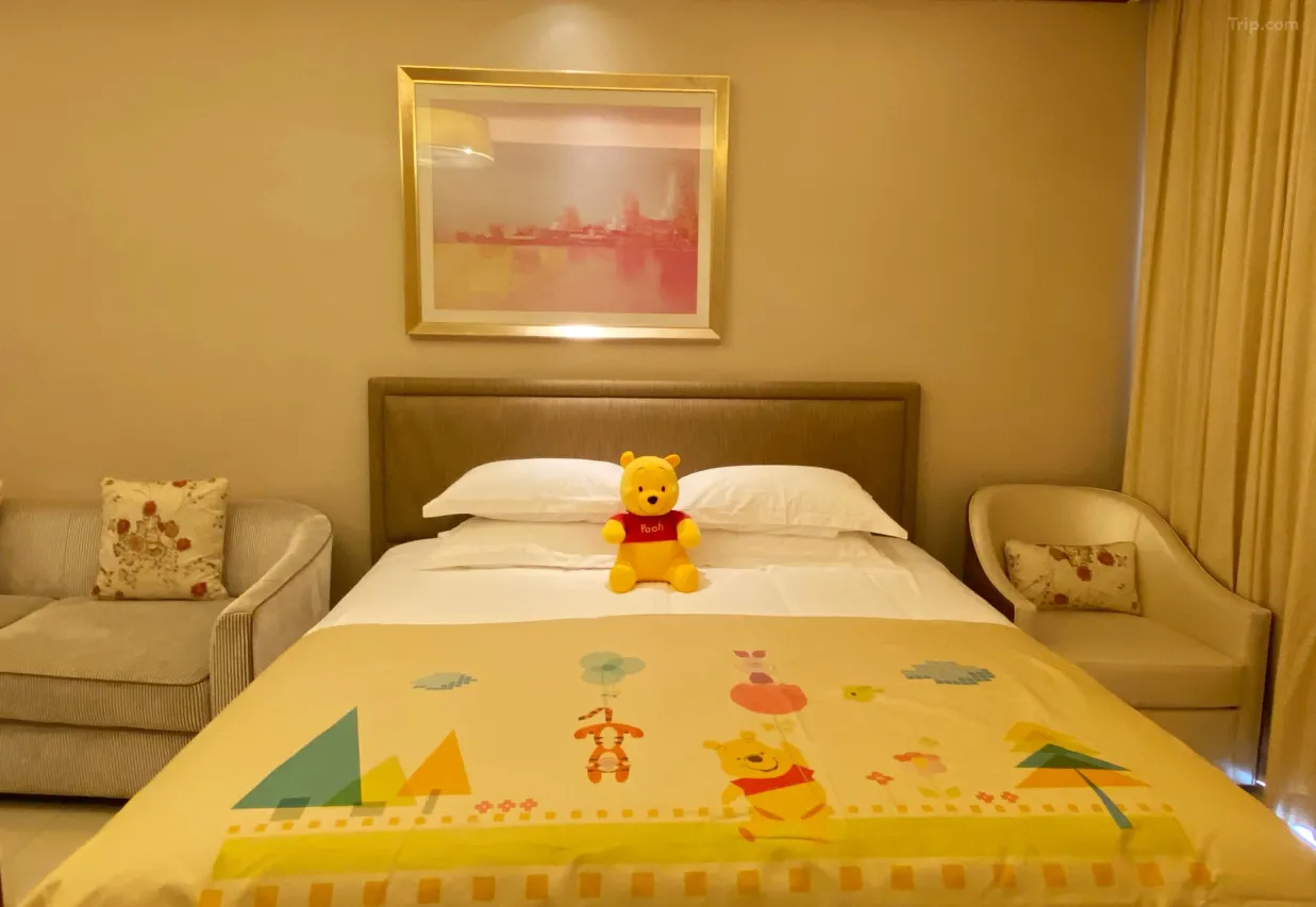 A bedroom with a bed in the center, topped with a Winnie the Pooh blanket and a Pooh plush toy. On either side of the bed are armchairs with floral pillows. There is a framed picture hanging above the bed and curtains on the side.