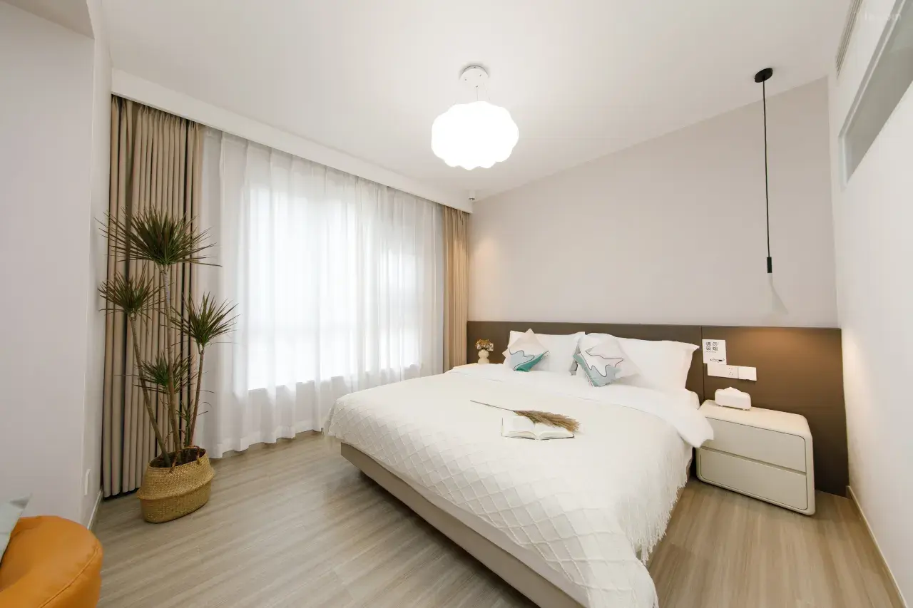 Modern hotel room with a large bed, white bedding, pillows, book, bedside table, plant, and window with curtains.