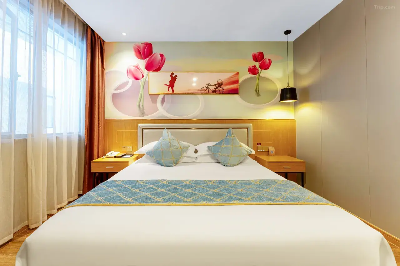 A neatly made hotel bed with light blue and beige pillows, a bed runner, and a beige headboard. The room has a large window with sheer white curtains and brown drapes, wall art with pink tulips, a silhouette of a person running, and a bicycle. There 