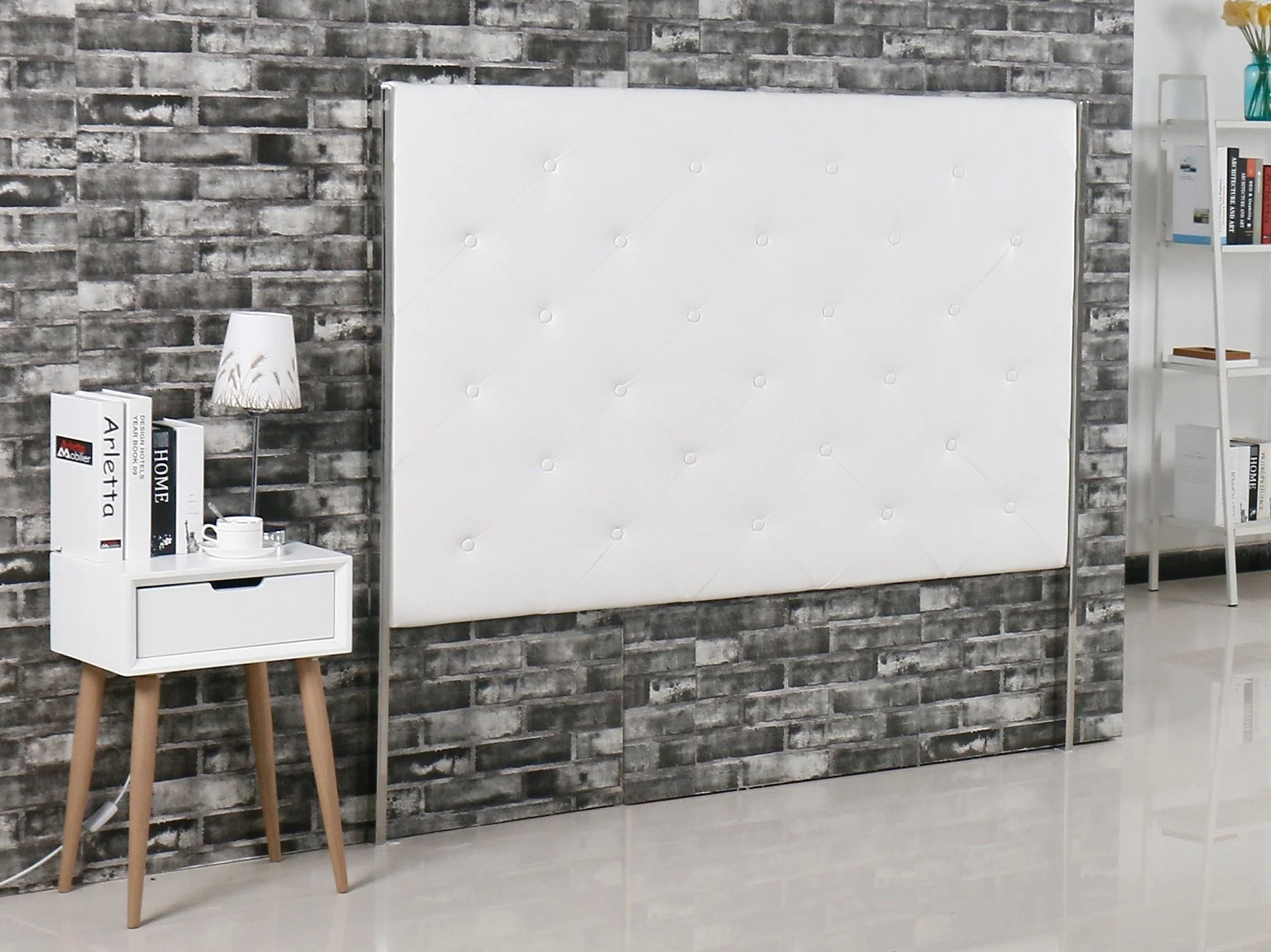 Interior scene with a white upholstered headboard against a gray brick wall, a white nightstand with books and a lamp, a coffee cup, and a small shelf with books and decorative items.