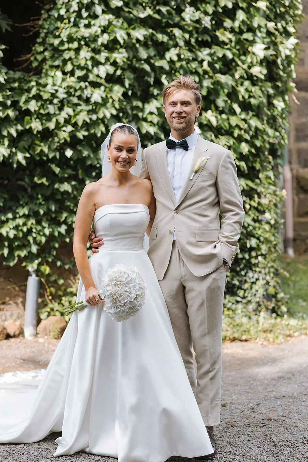Happy bride and groom in wedding attire posing outdoors in front of green ivy-covered wall.