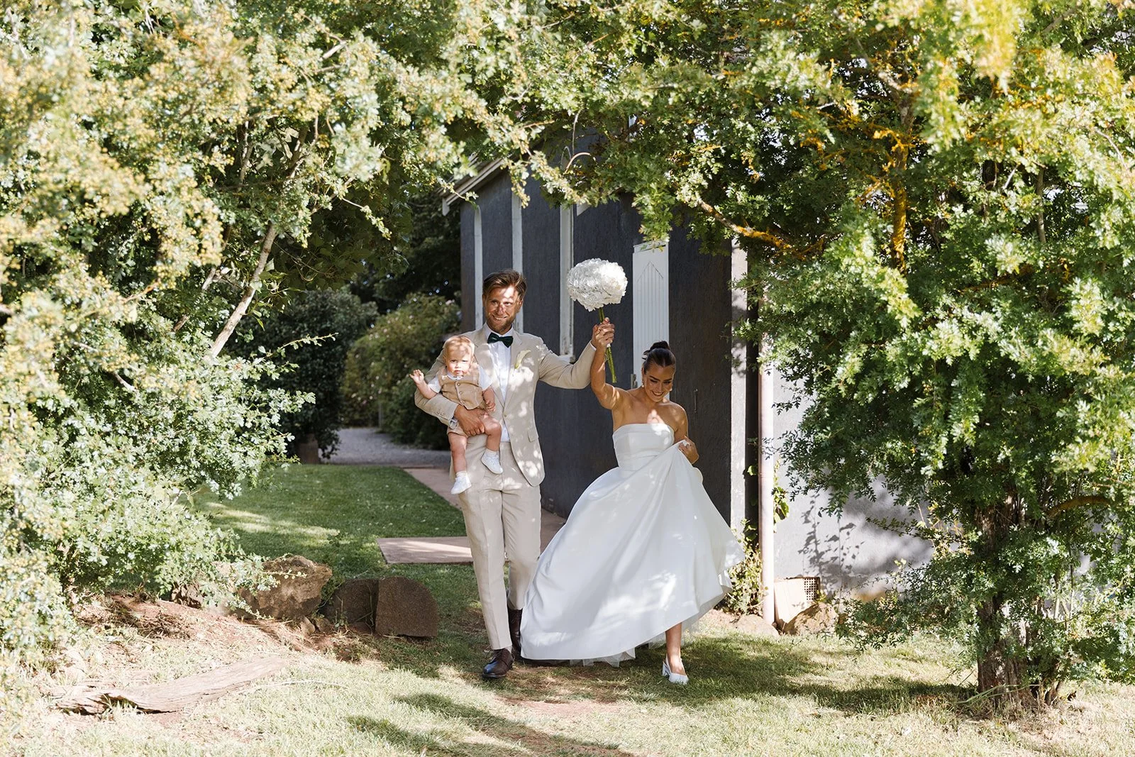 Wedding couple walking outdoors under trees, the bride is holding a bouquet up, and the groom carries a child, all smiling and dressed in wedding attire.