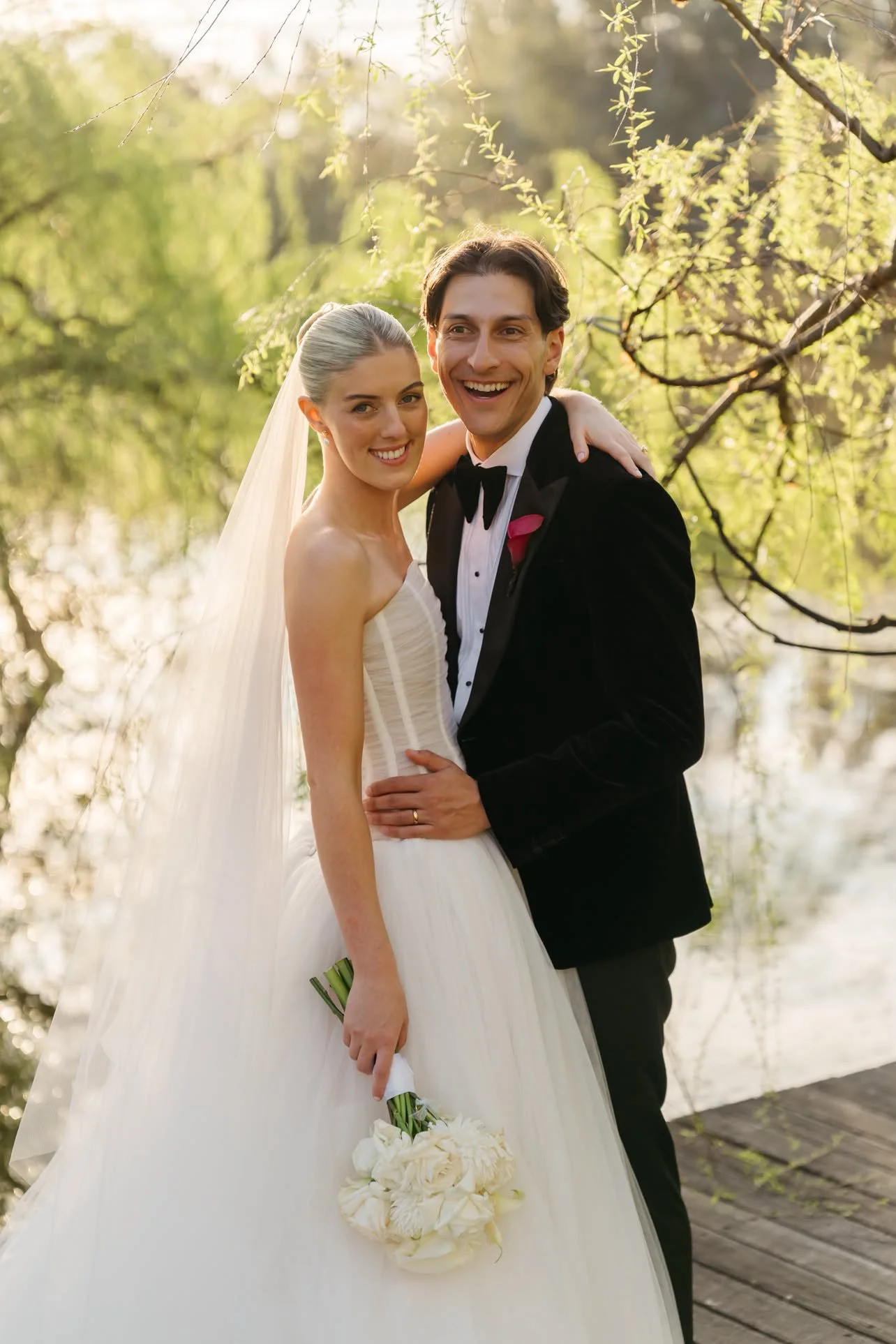 A wedding couple outdoors, the bride holding a bouquet of white flowers, and the groom in a tuxedo, both smiling.