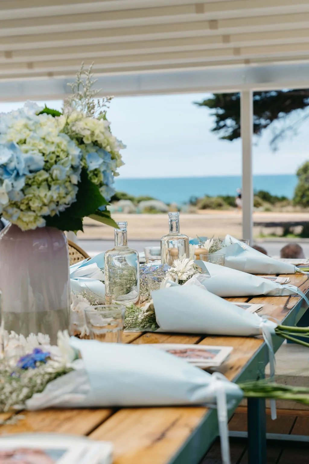 A table set for an outdoor event near the beach, decorated with floral arrangements, glass bottles, and folded napkins.