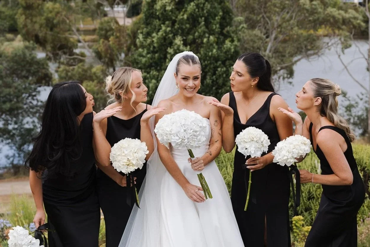 A bride in a white wedding gown holding a bouquet of white flowers, smiling, with four women dressed in black dresses, each holding a white flower bouquet, outdoor setting near a lake and trees.