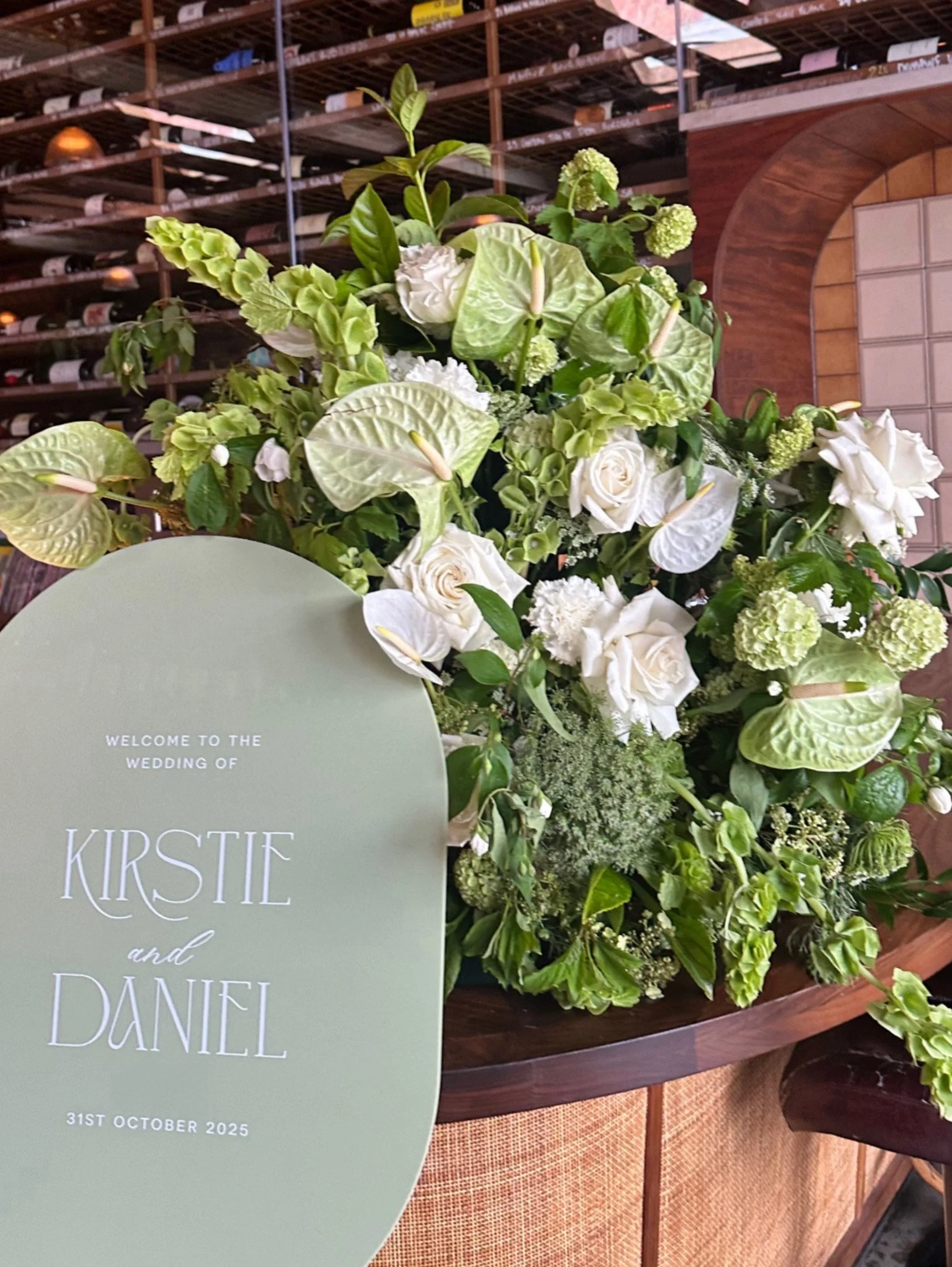 Close-up of a wedding welcome sign with the names Kirstie and Daniel, surrounded by a lush arrangement of white and green flowers, including roses, anthuriums, and hydrangeas, set on a wooden table.