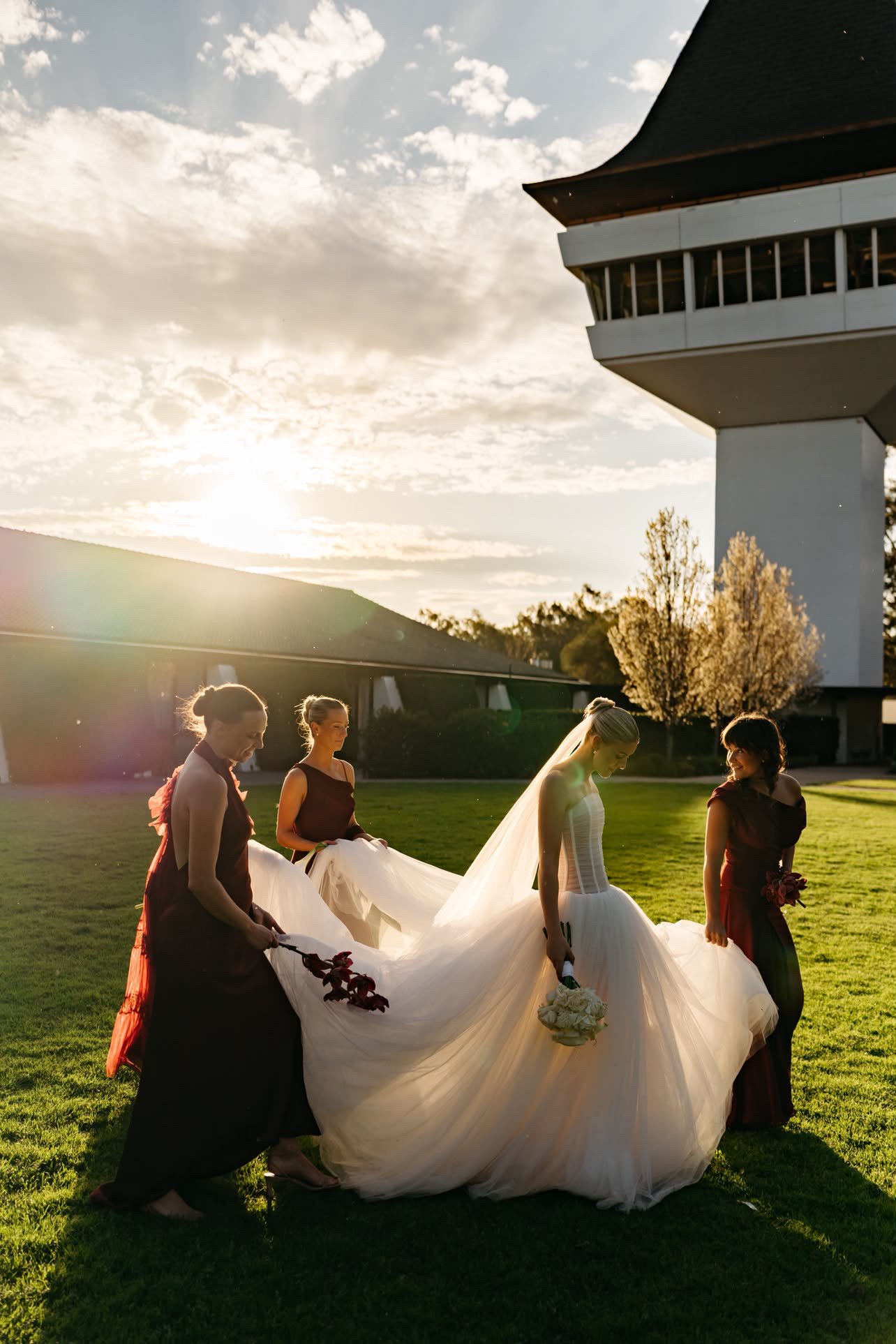 A bride and bridesmaids outdoors during sunset, with the bride in a white wedding dress and veil, holding a bouquet, and the bridesmaids in dark dresses holding the train of the dress on a grassy area with a modern building and trees in the background.