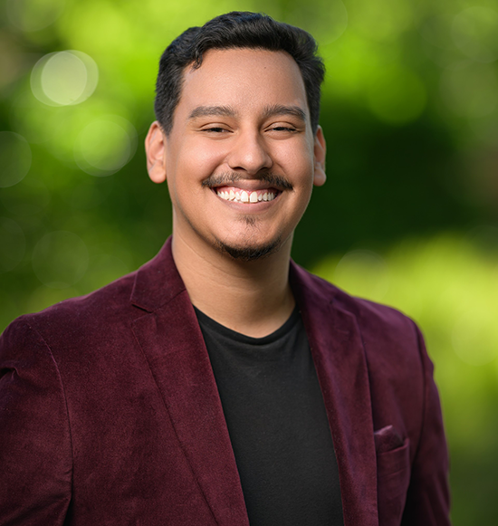 A young man smiling outdoors with a blurred green background, wearing a maroon blazer over a black shirt.