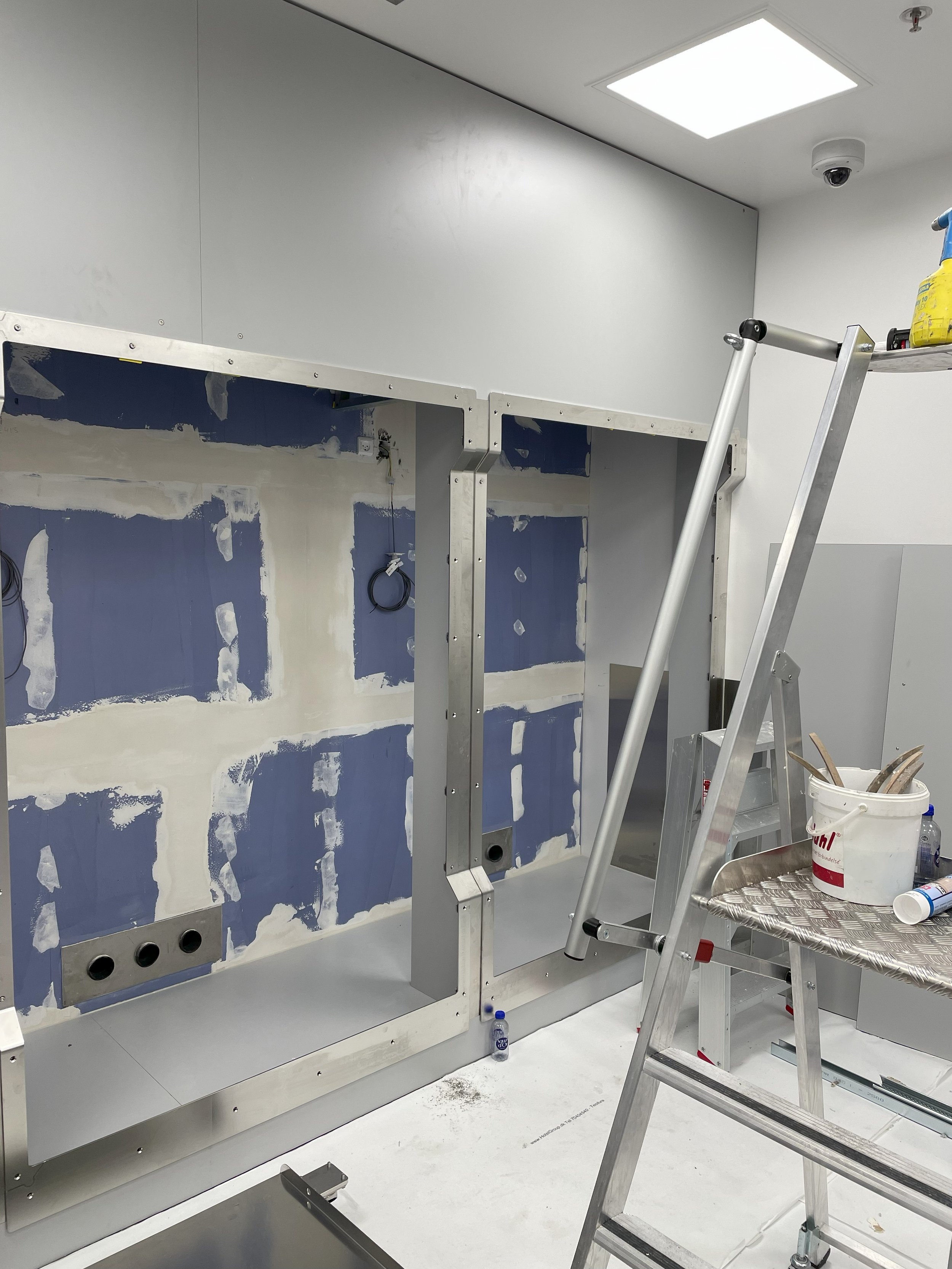 Construction site with partially assembled metal framework, unfinished blue drywall, electrical wiring, and tools including a ladder and a bucket.