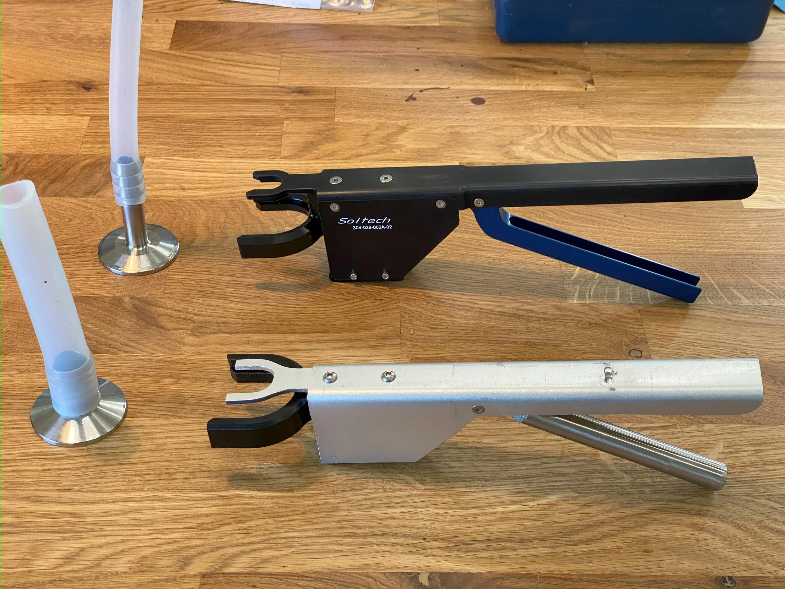 Two clamper tools, one black and one silver, placed on a wooden surface. The black tool has the brand name 'Soltech' and some numbers, the silver one has a curved metal handle.