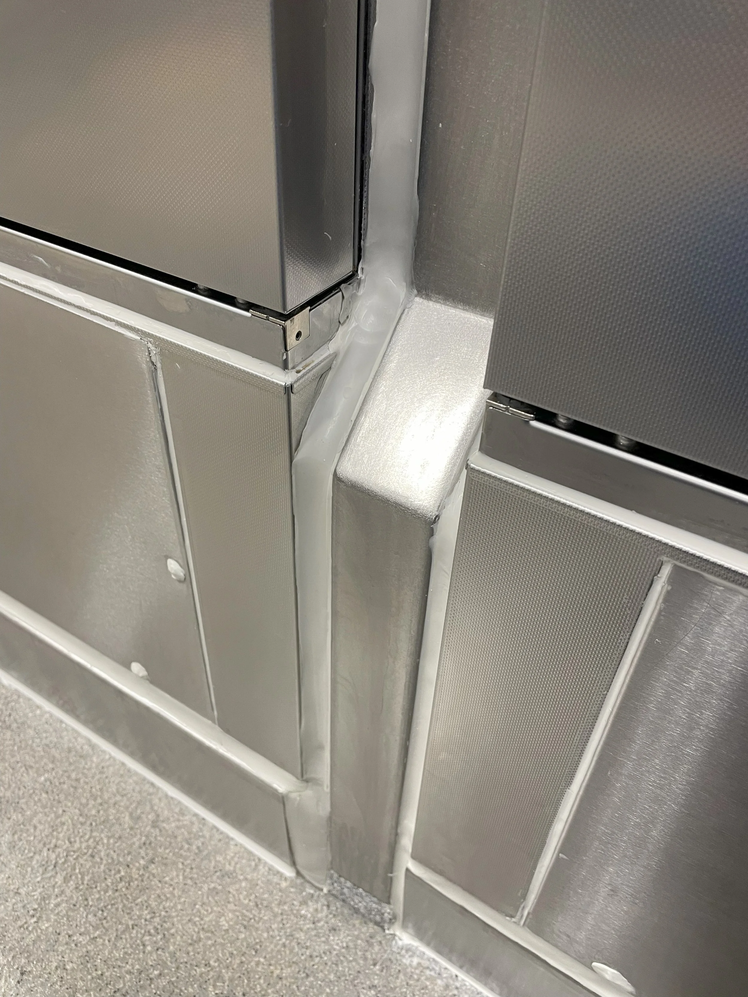Close-up of a corner in a kitchen or industrial area showing stainless steel panels and a caulked seam.