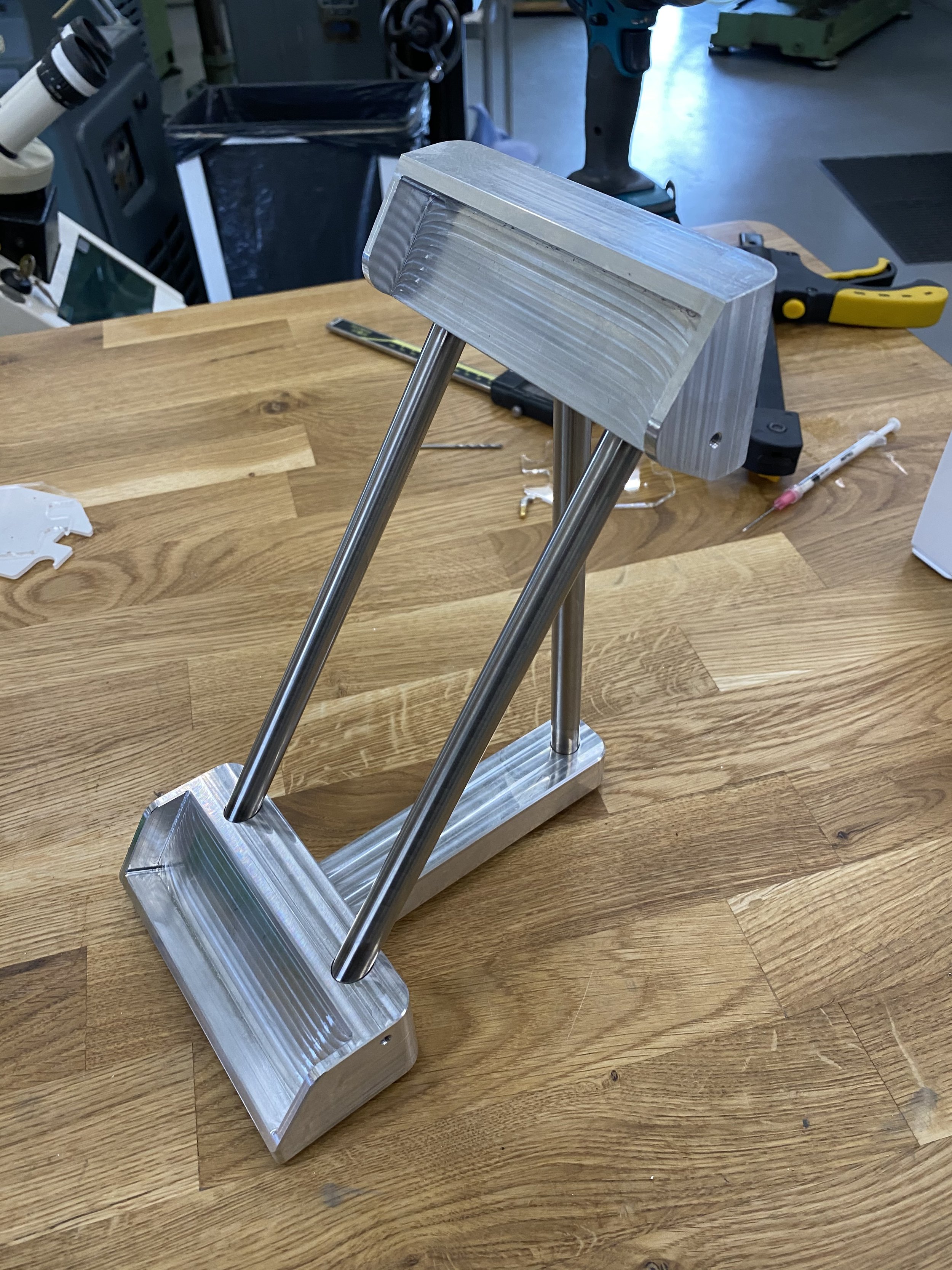A metallic stand with two angled rods supporting a flat top and bottom base, positioned on a wooden workbench.