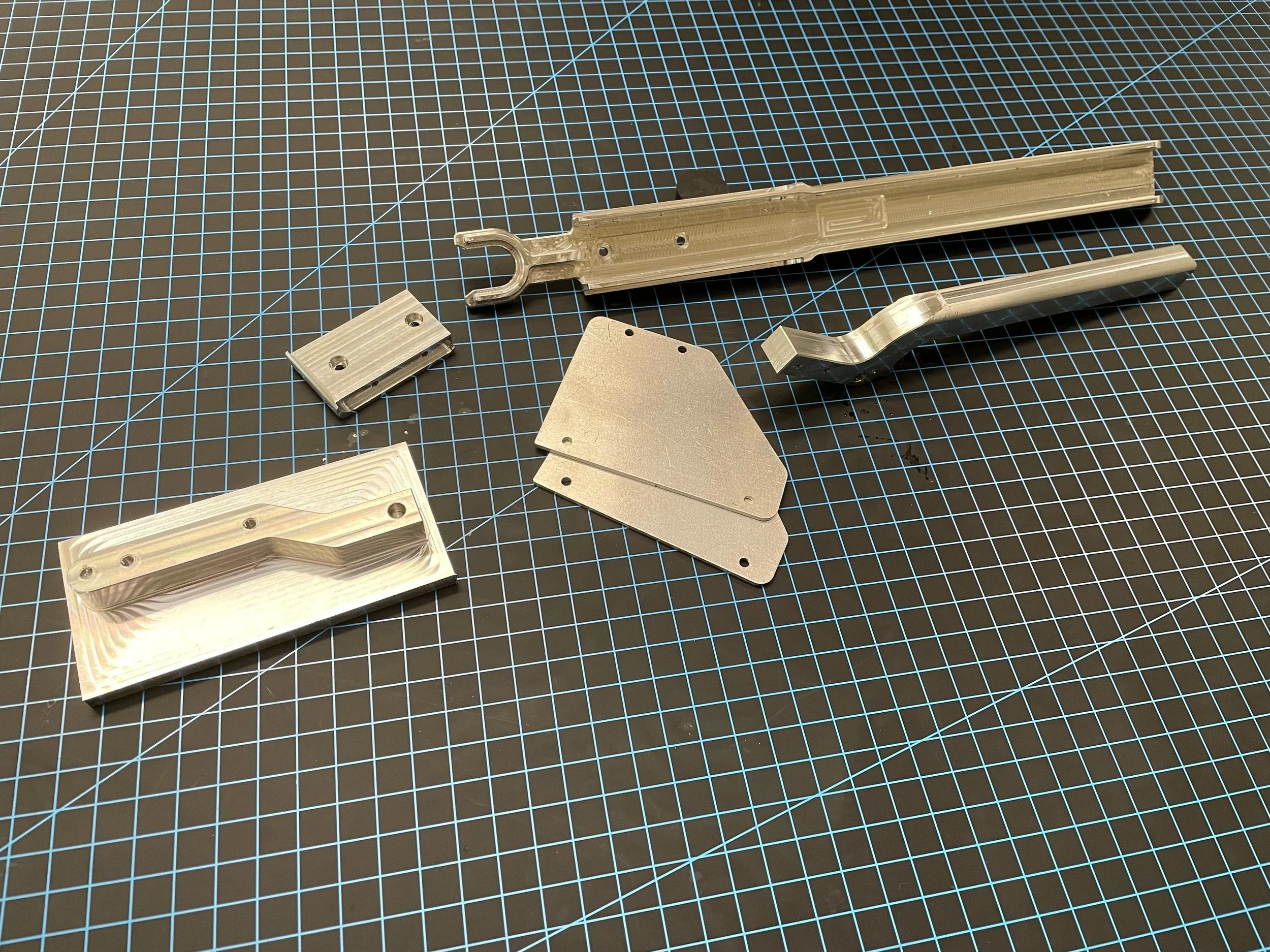 Various silver metal parts and machined components laid out on a black workshop mat with blue grid lines.