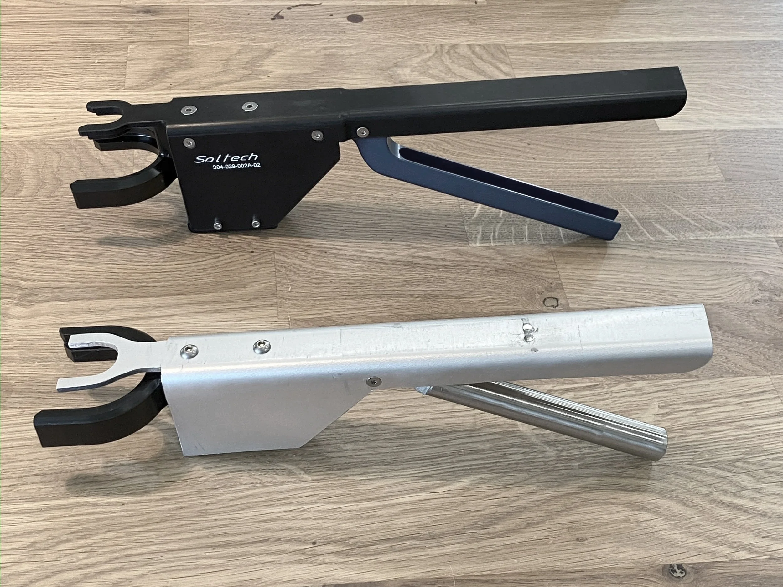 Two metal automotive tow hooks, one black and one silver, are placed on a wooden floor, with the black hook positioned above the silver hook.