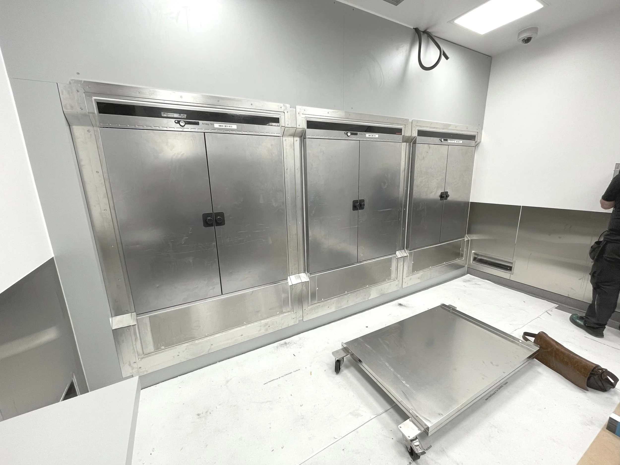 A room with stainless steel doors and panels, with a wheeled stainless steel platform and a brown cylindrical object on the floor.