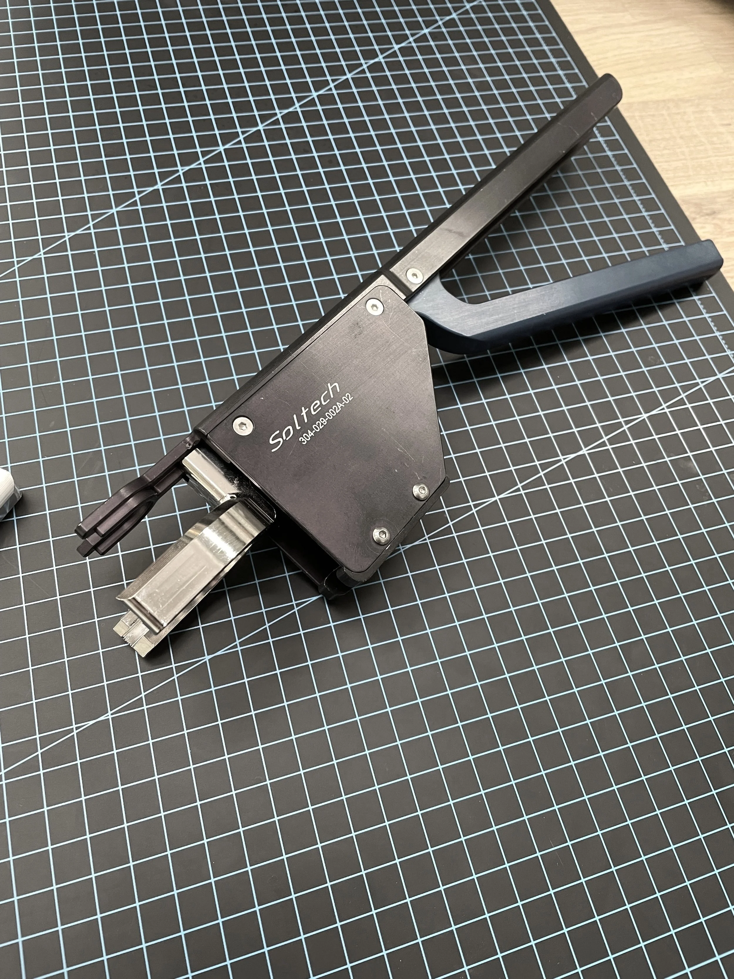 A black and silver mechanical device with the brand name Soltech and serial number, resting on a black cutting mat with a grid pattern.