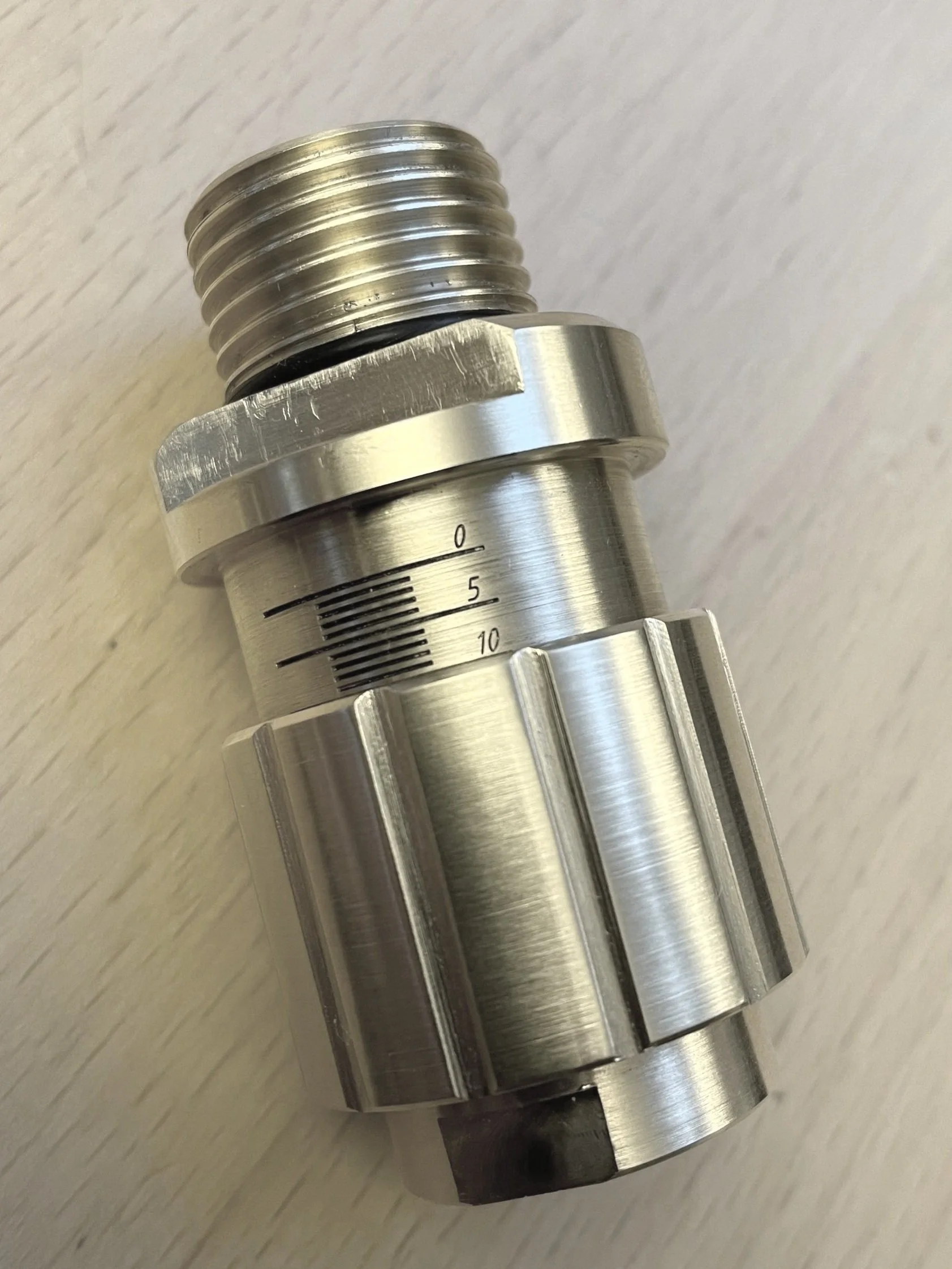 A close-up of a metallic industrial component, cylindrical in shape with a threaded top, a scale with black lines, and grooves in the body.