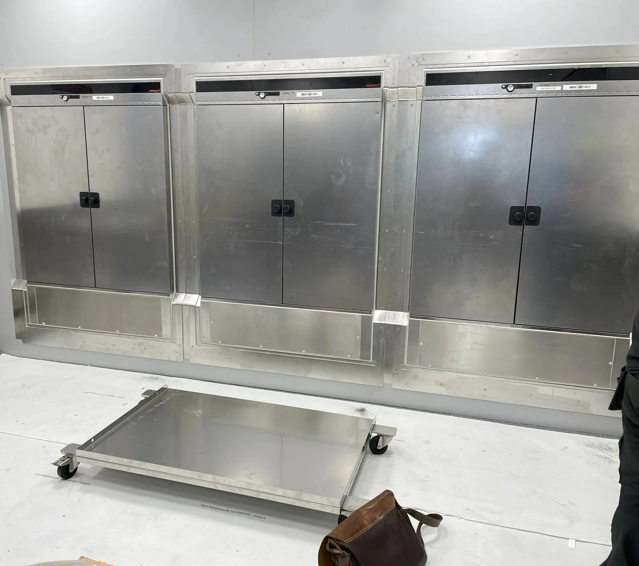 Industrial stainless steel electrical equipment cabinets behind a metal platform on wheels and a brown leather bag on white flooring.