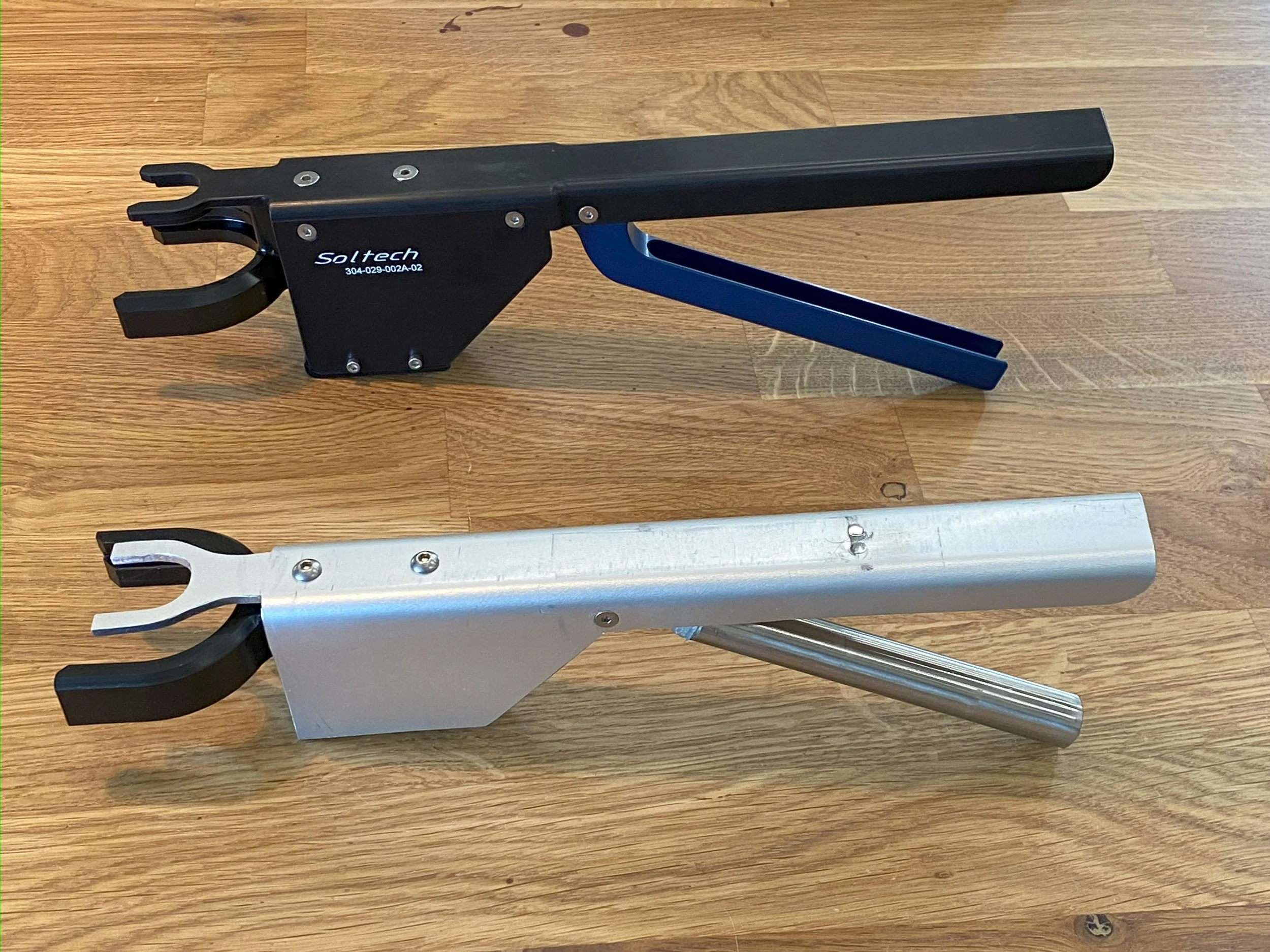 Two metal tools with open-ended prongs at one end, one black and one silver, resting on a wooden surface.