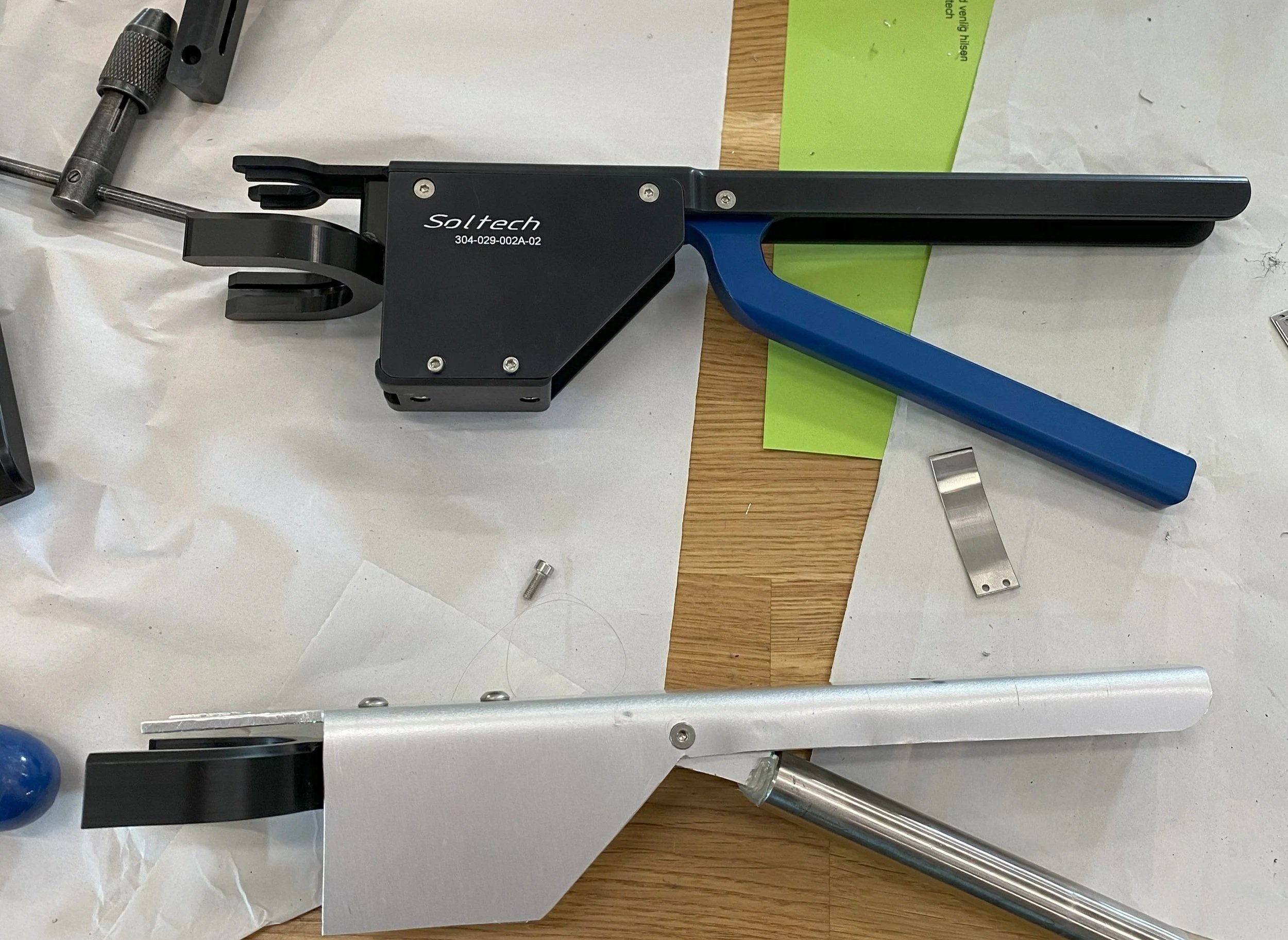 Two metal frame tools with black and blue handles, resting on paper and a wooden surface, with small metal parts nearby.