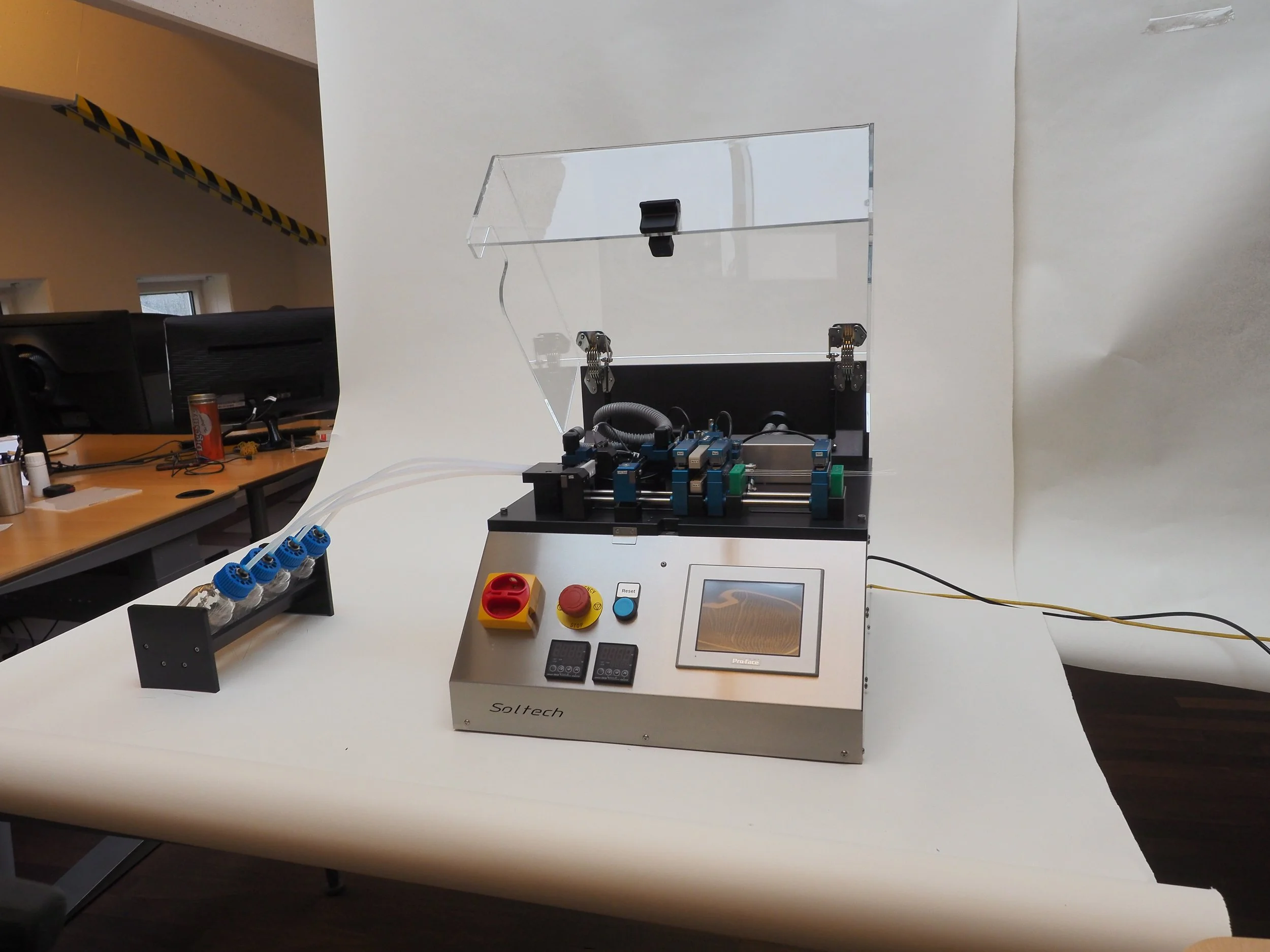 A piece of laboratory equipment with a touchscreen display, emergency stop button, and multiple small glass bottles connected by tubes.