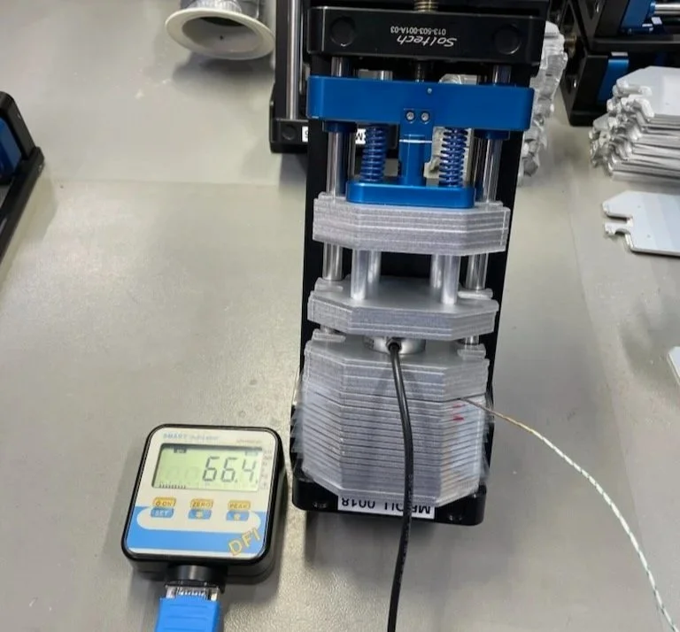 A digital meter displaying a reading of 66.4 connected to a machine with stacked gray layers, blue and black components, and a cable attached, on a gray work surface.