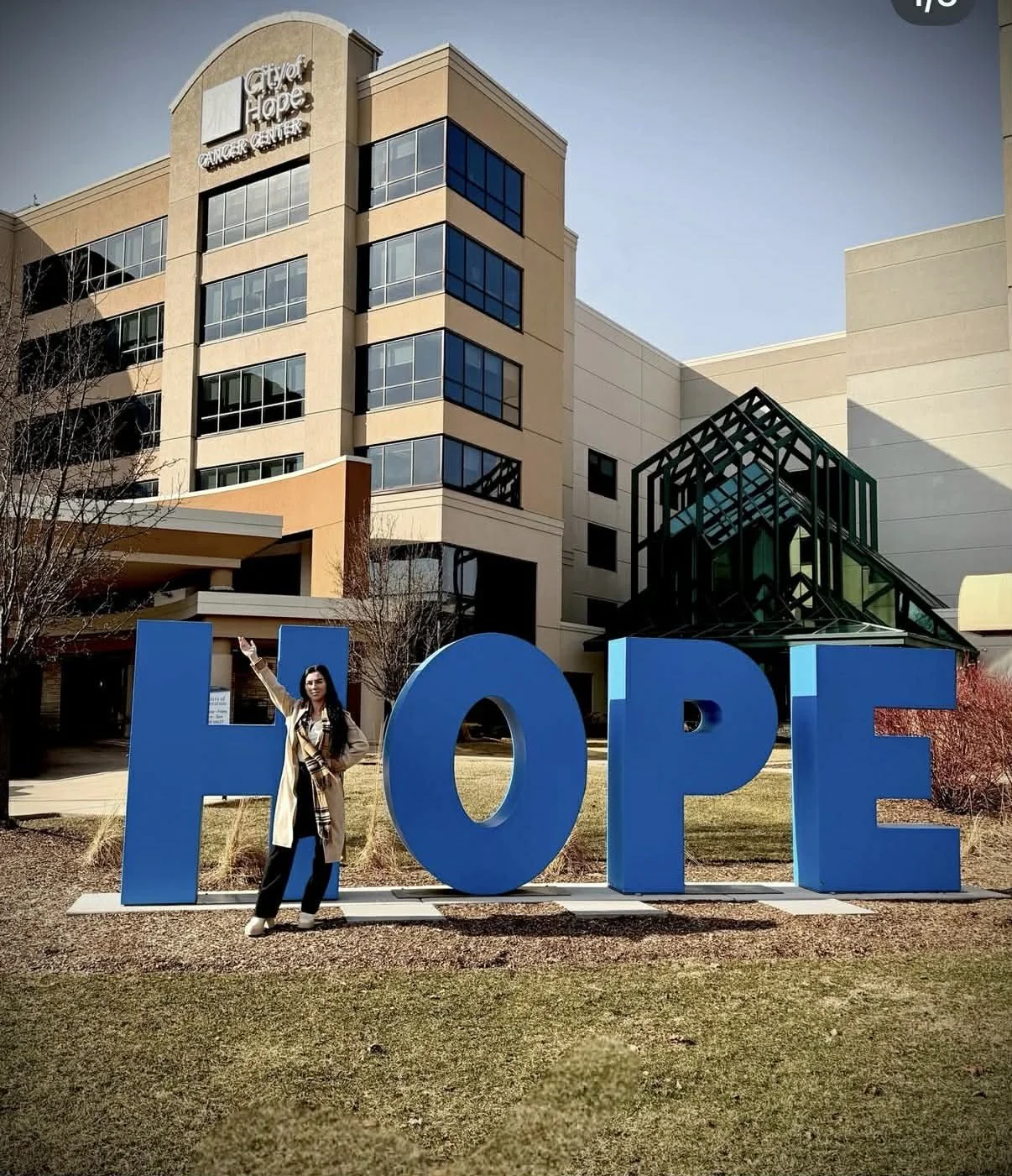A woman standing next to a large blue sign that spells 'HOPE' outside a modern building with the sign 'City of Hope Cancer Center' on it.