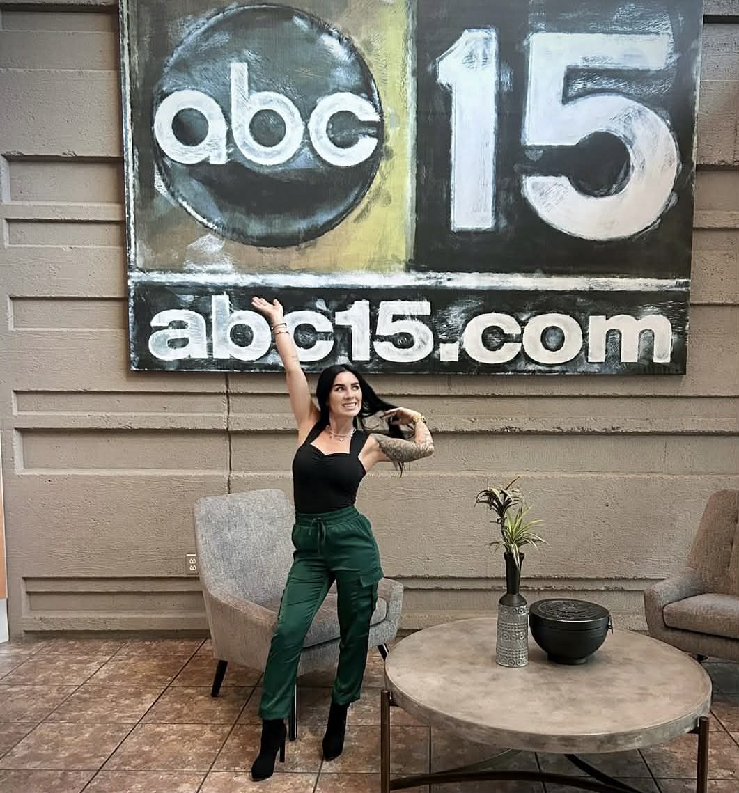 A woman with long dark hair, wearing a black top and green pants, poses with one arm raised and smiling in front of a large sign that reads 'abc 15' and 'abc15.com,' inside a room with tan walls, a round table with a decorative bowl and a potted plant, and a gray armchair.