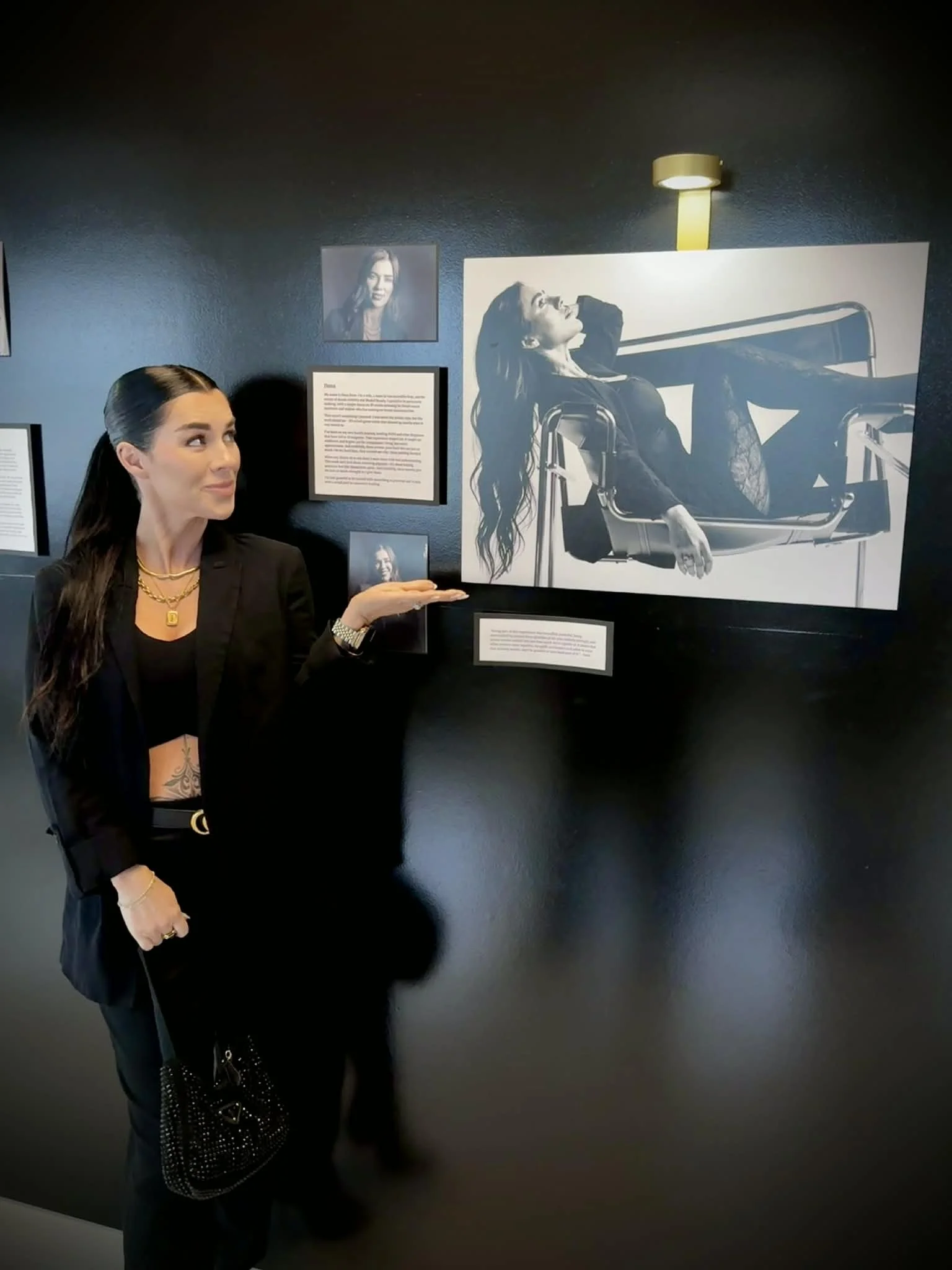 A woman in black outfit and gold jewelry pointing at a large black-and-white photograph of a woman reclining in a modern lounge chair at an art exhibit.