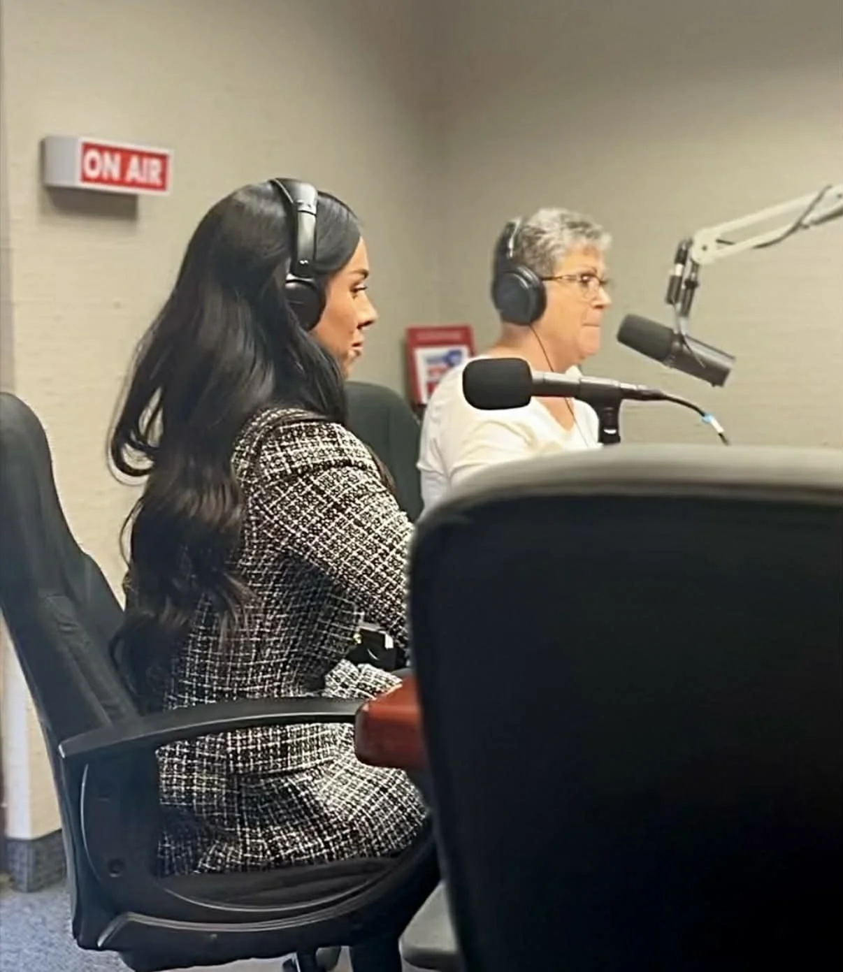 Two people sitting in a recording studio wearing headphones, speaking into microphones, with an 'ON AIR' sign in the background.