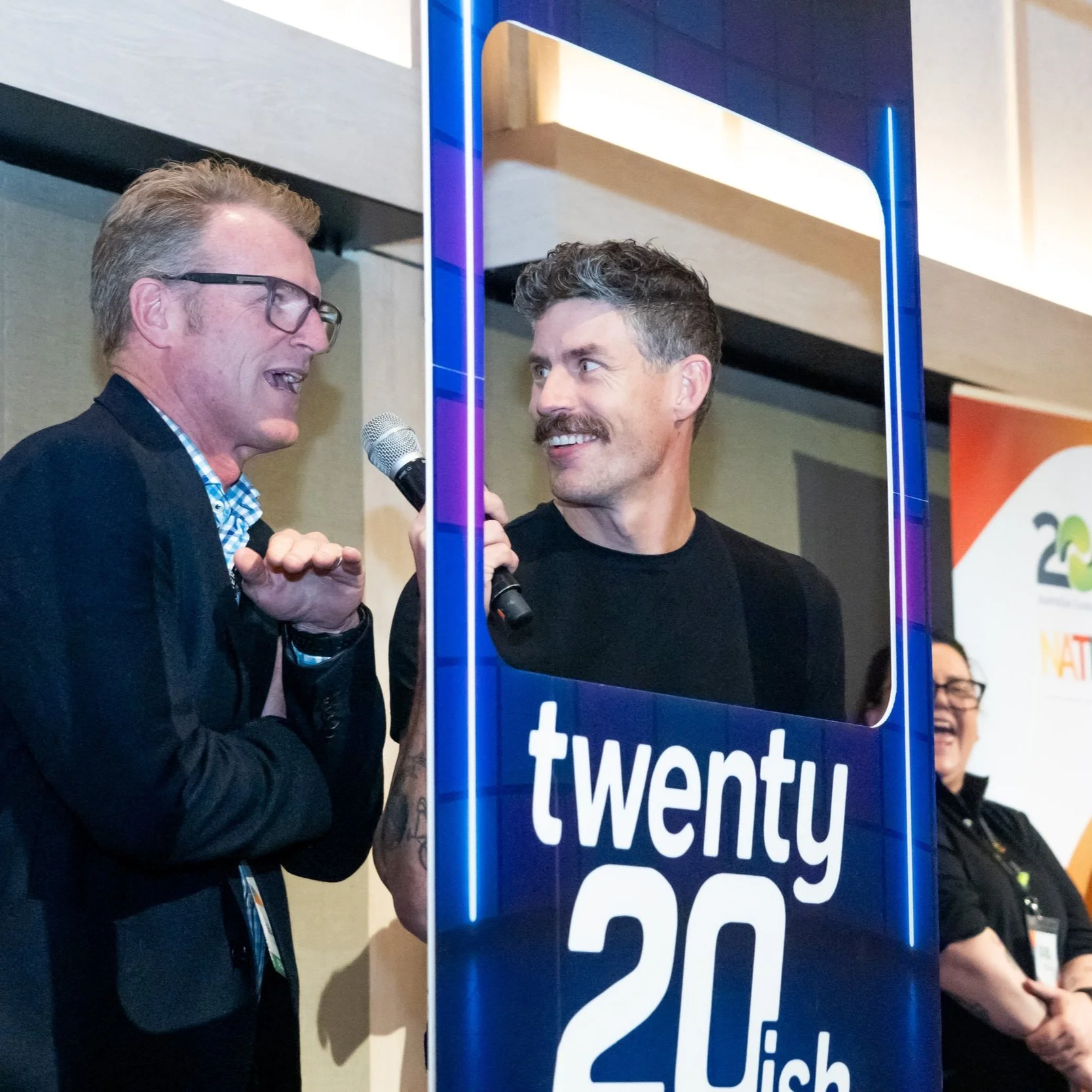 Luke Jamieson smiling and talking at a conference, one holding a microphone, framed by a blue sign reading 'twenty20ish'.