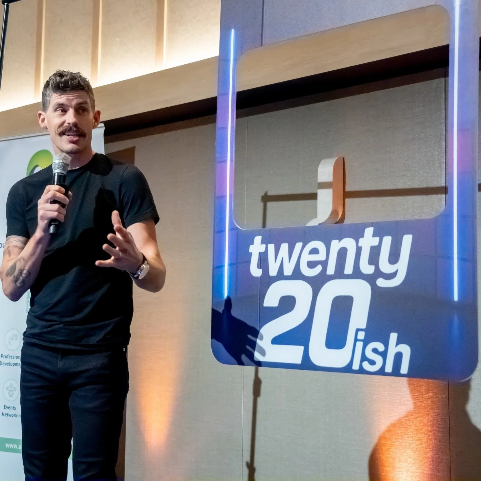 Luke Jamieson with a microphone speaking at a Twenty20ish event, standing next to a sign that says 'twenty20ish' with a stylized logo.
