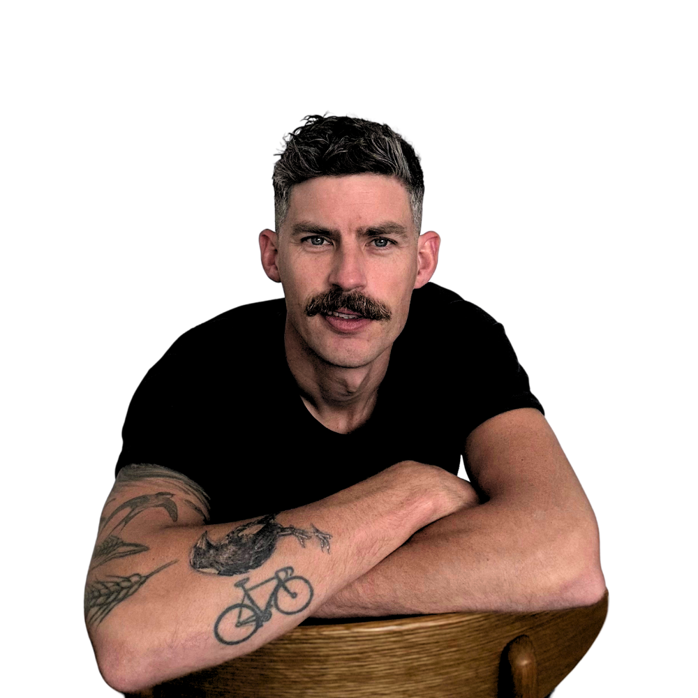Luke Jamieson, A handsome man with a moustache and short, dark hair, wearing a black t-shirt, resting his forearms on a wooden surface, with tattoos on his left arm, against a white background. Twenty20ish.