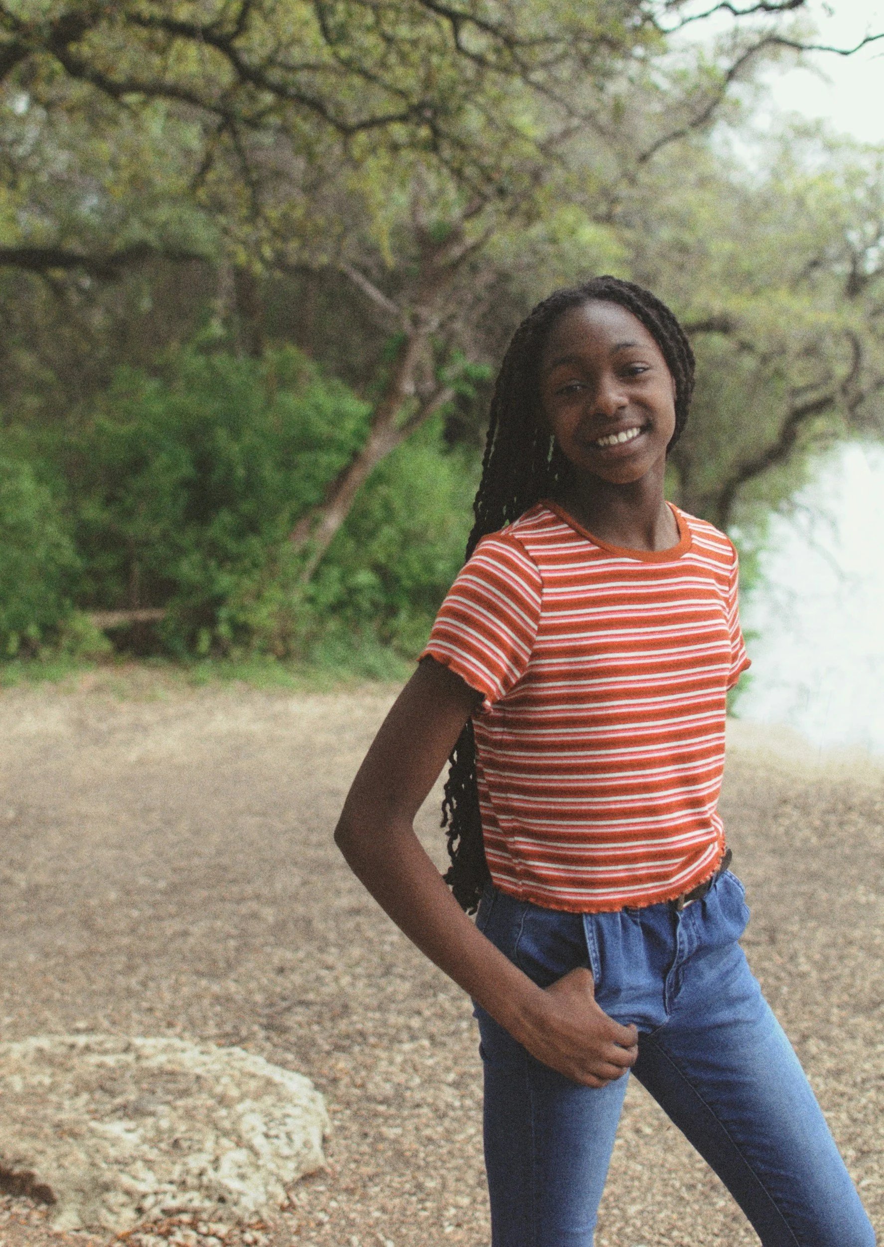 a young black teen wears blue jeans and a red and white striped t-shirt. They have their right thumb in their right front jeans picket and they are smiling with teeth showing. They are outside in nature with trees and shrubs behind them.