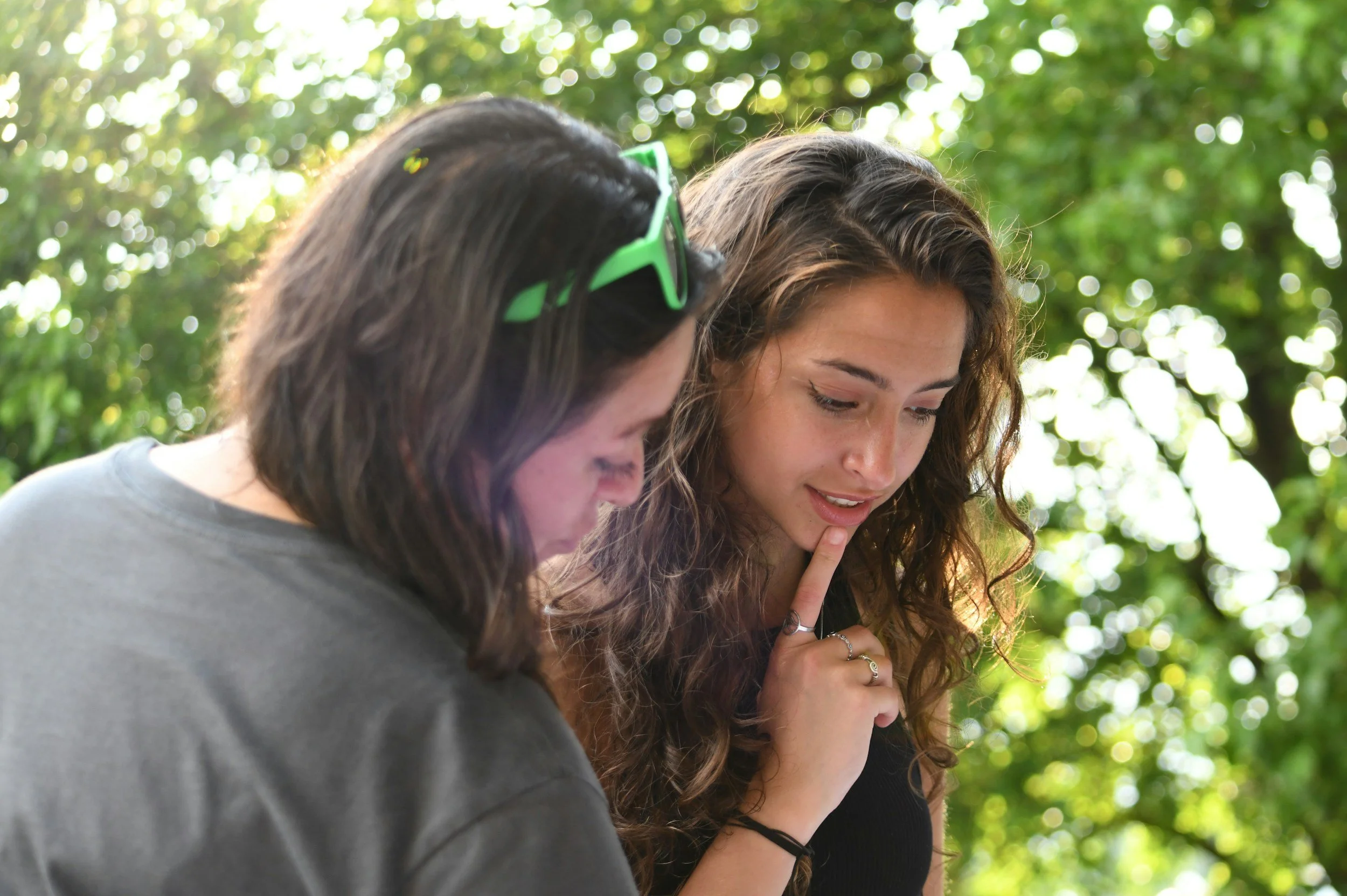 Two girls are outside by trees looking down at something. They both have dark hair, and one of them has curly hair and has a finger on her chin. She has a ring on her pointer finger and a black bracelet. The other person has green sunglasses.