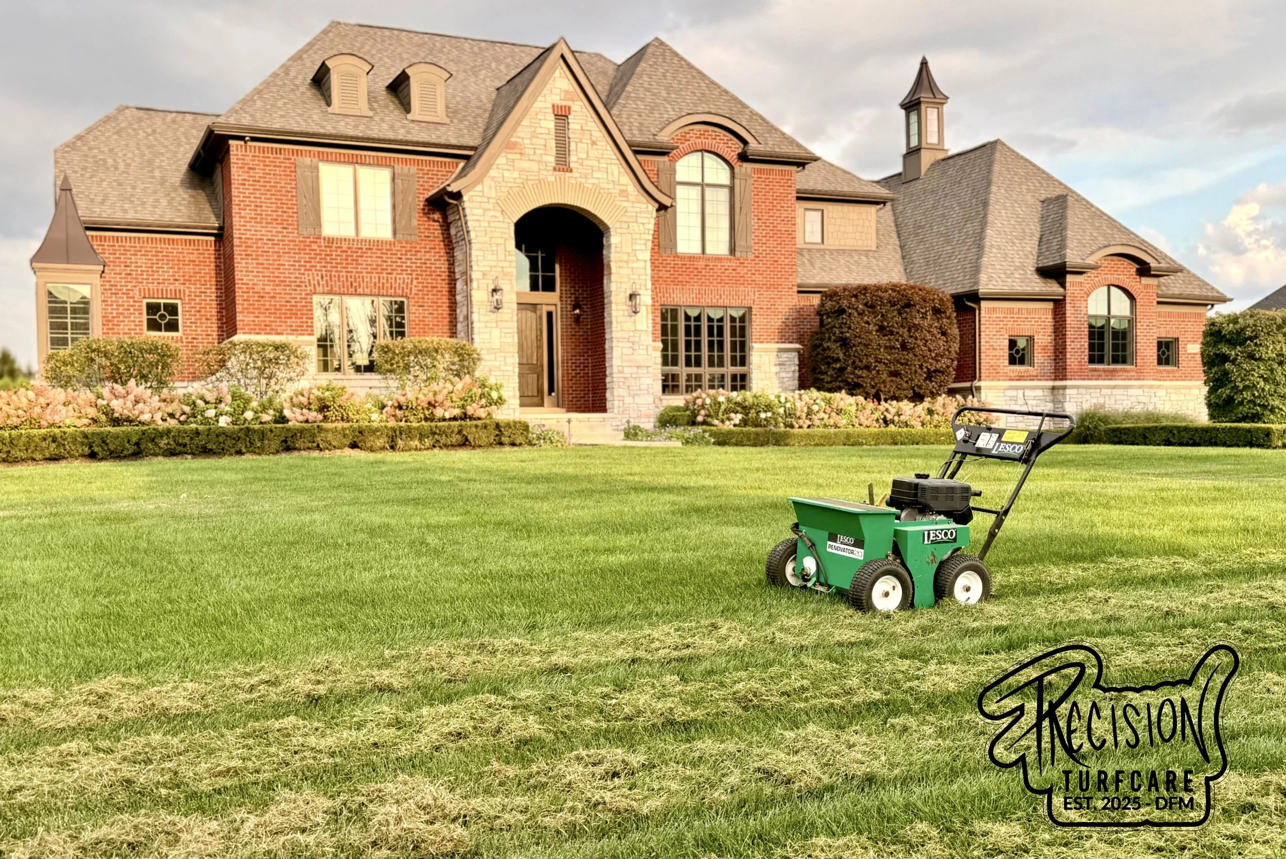 A large, luxurious brick house with a manicured lawn and a green turf care machine in the foreground.
