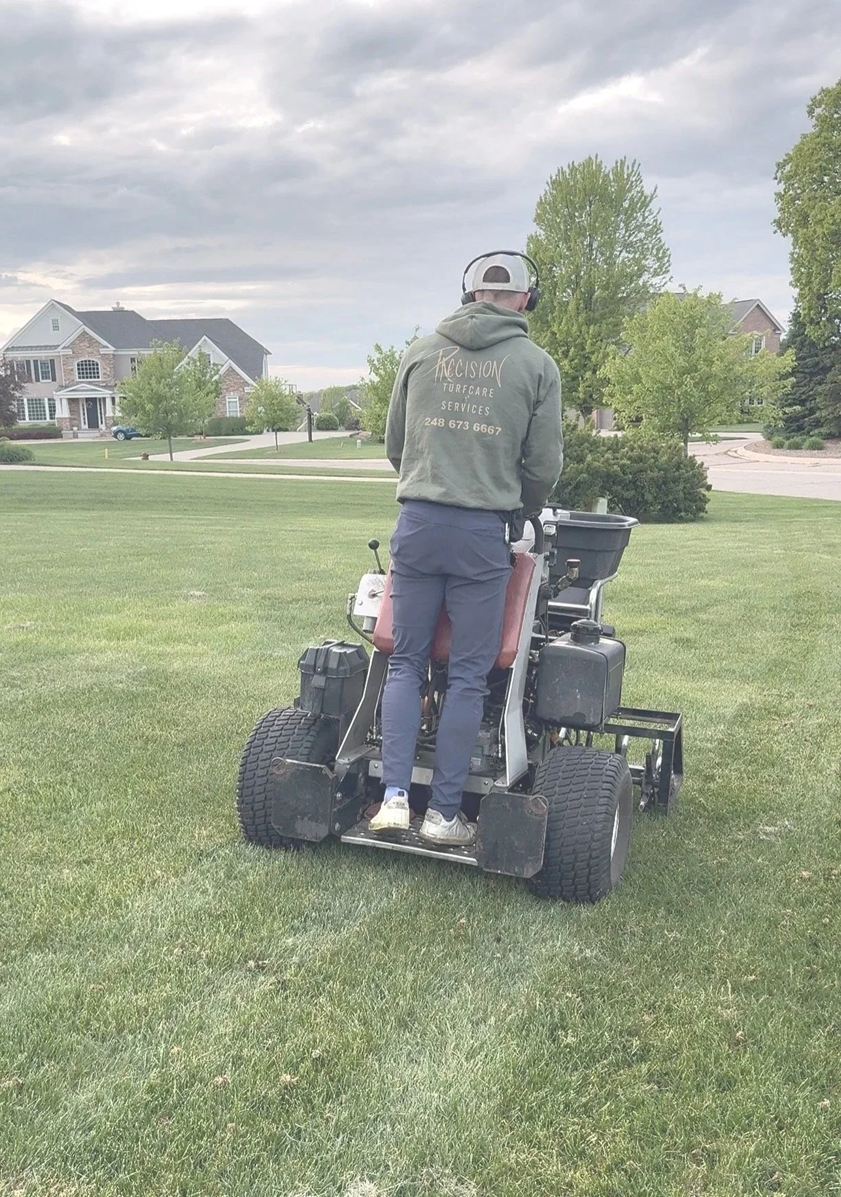 Man operating a turf care machine on a well-maintained lawn in a suburban neighborhood with houses and trees in the background.