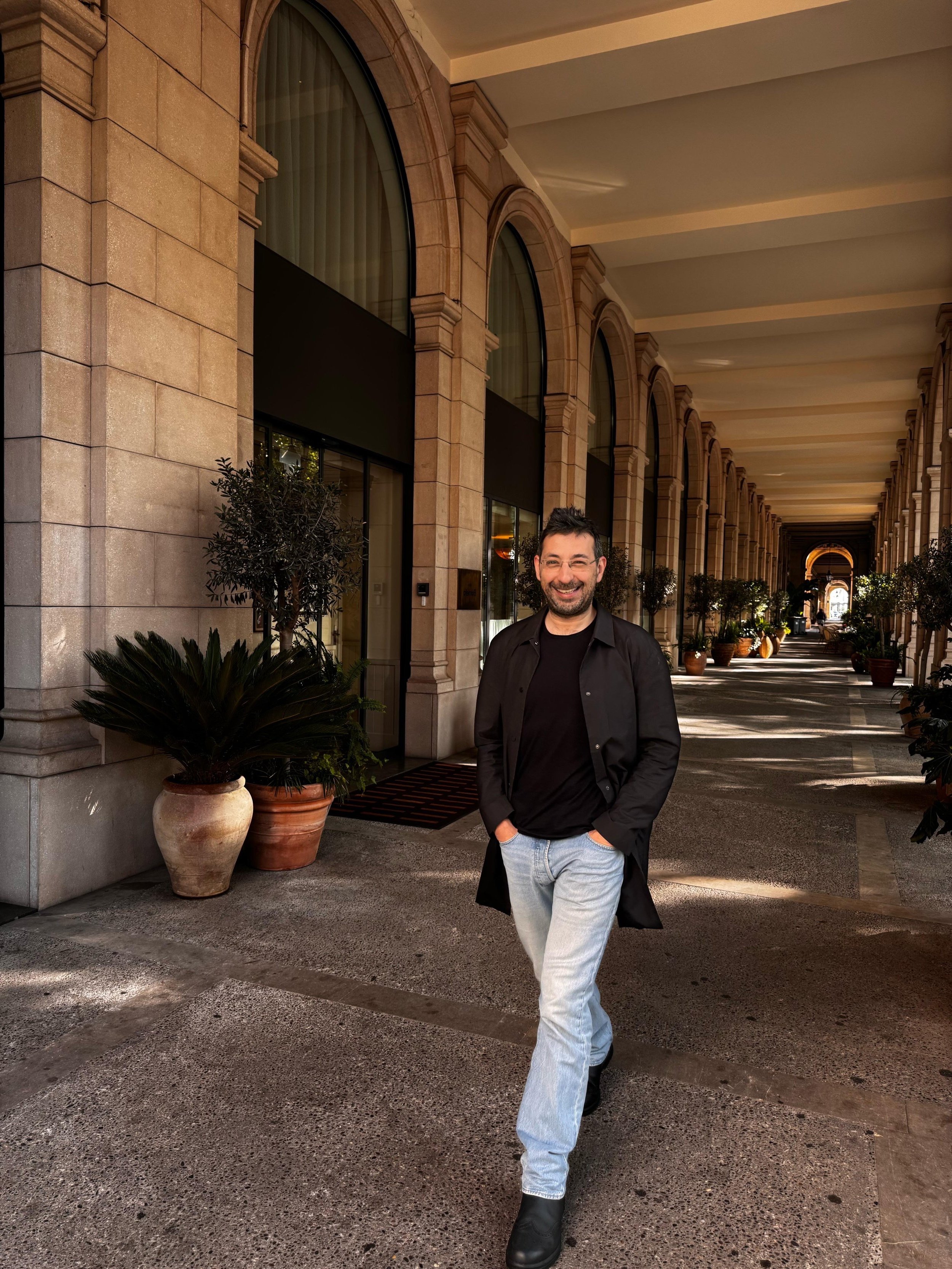 Man with black hair, beard, and glasses walking in a covered outdoor corridor with potted plants, wearing a black coat, black shirt, light blue jeans, and black shoes, smiling at the camera.